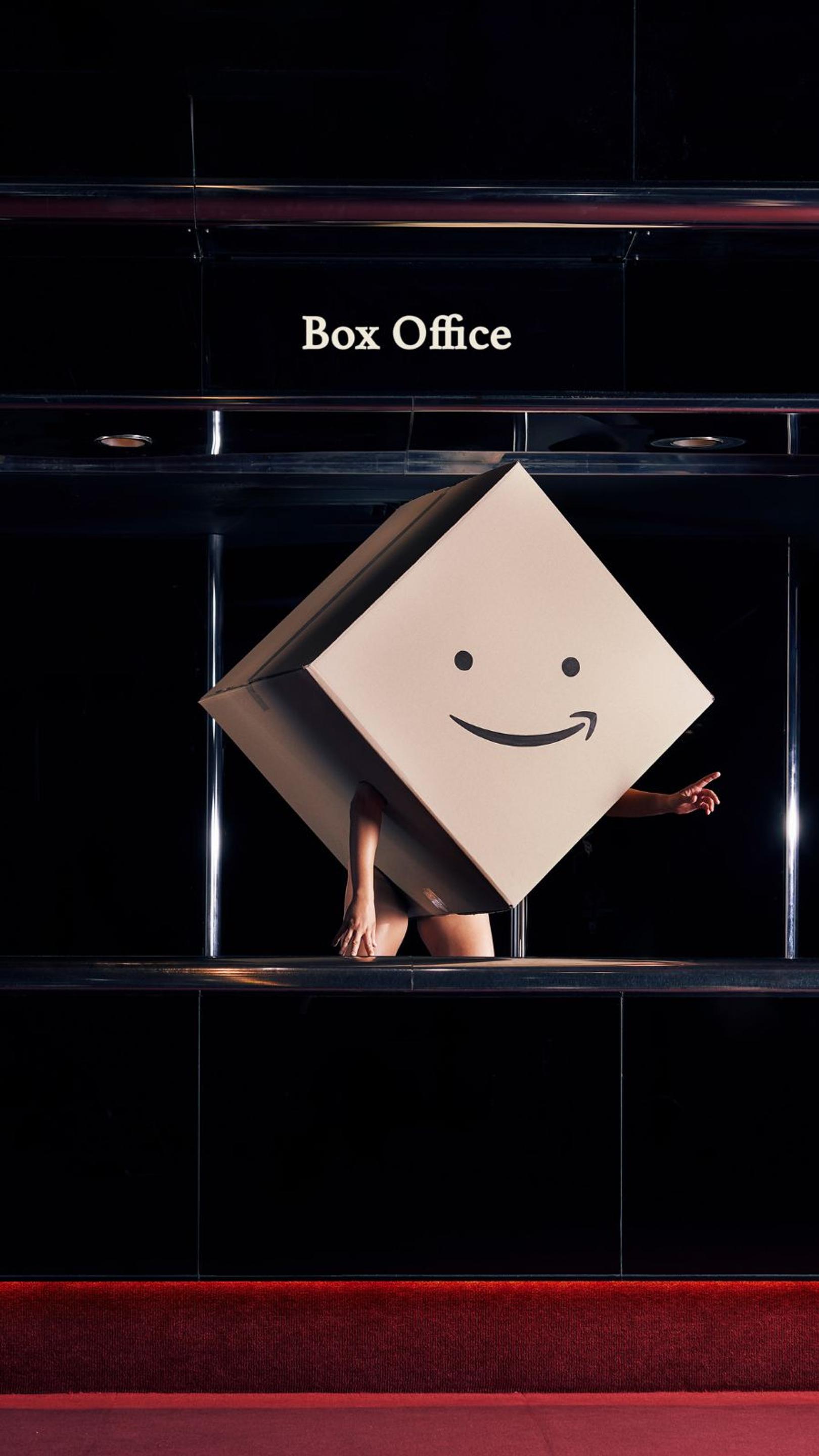 A person wearing a giant box, printed with a smile on the front, stands in a dim Box Office. 