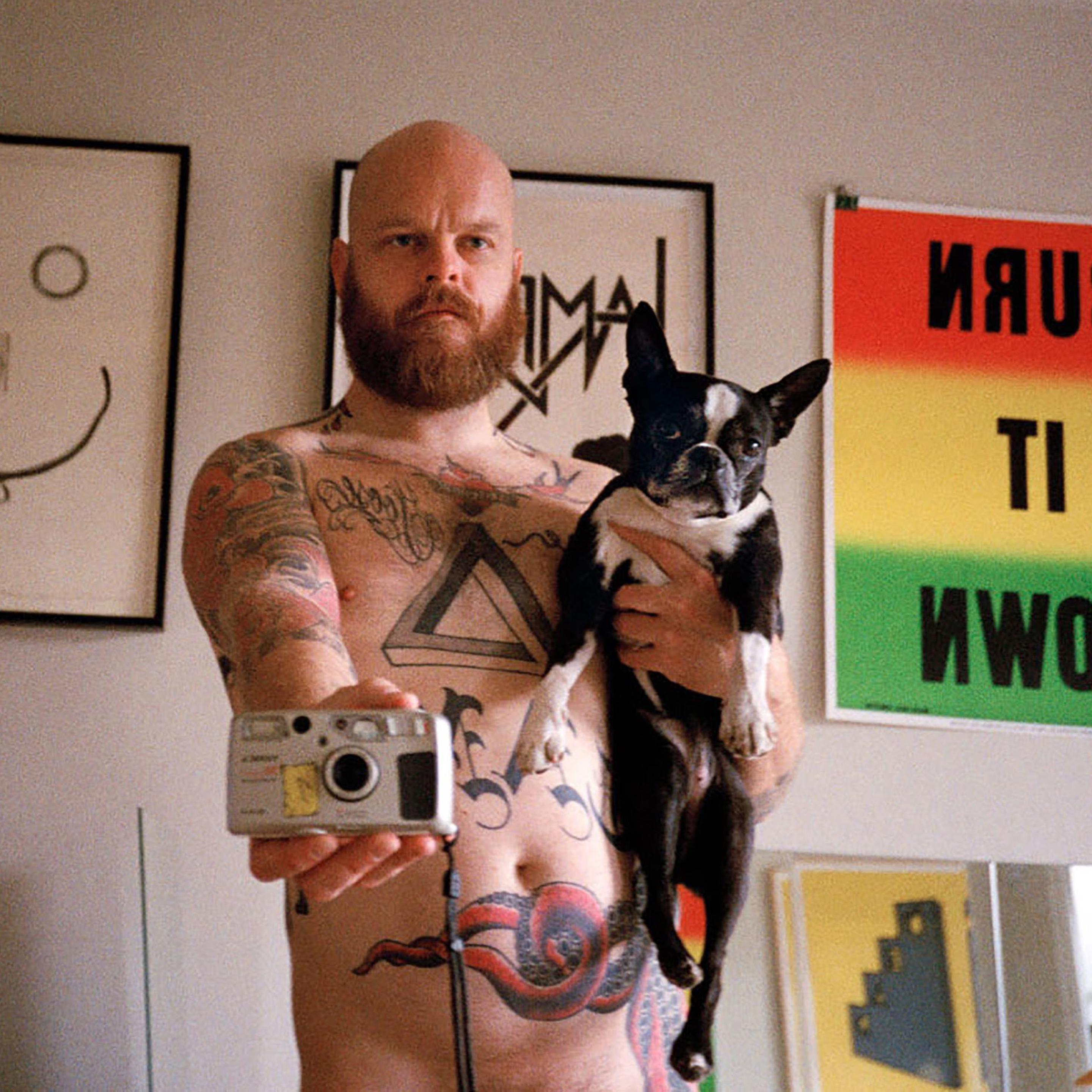 Shannon Michael Cane takes a nude selfie in a mirror, holding a french bulldog in one hand and the film camera in the other.