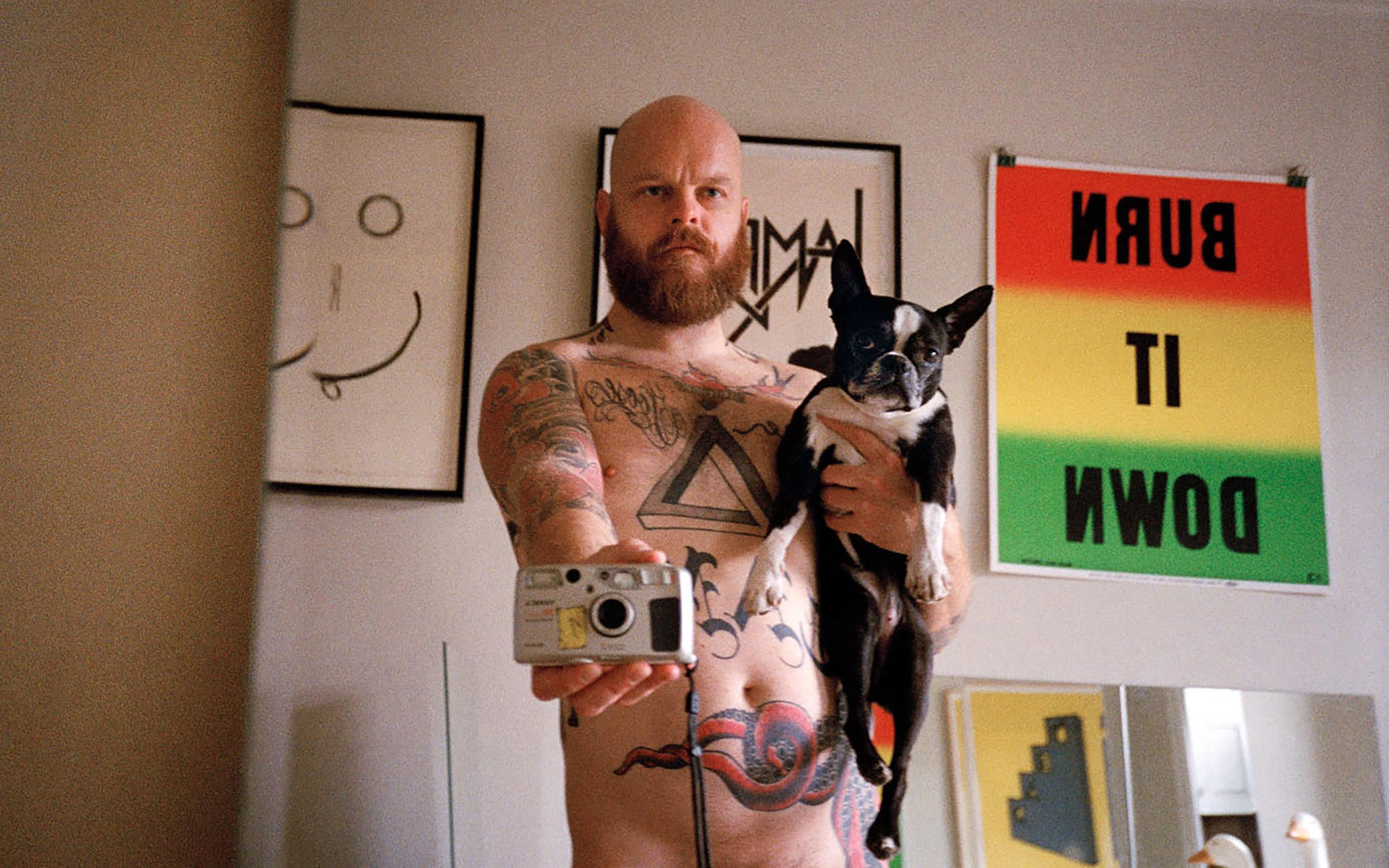 Shannon Michael Cane takes a nude selfie in a mirror, holding a french bulldog in one hand and the film camera in the other.