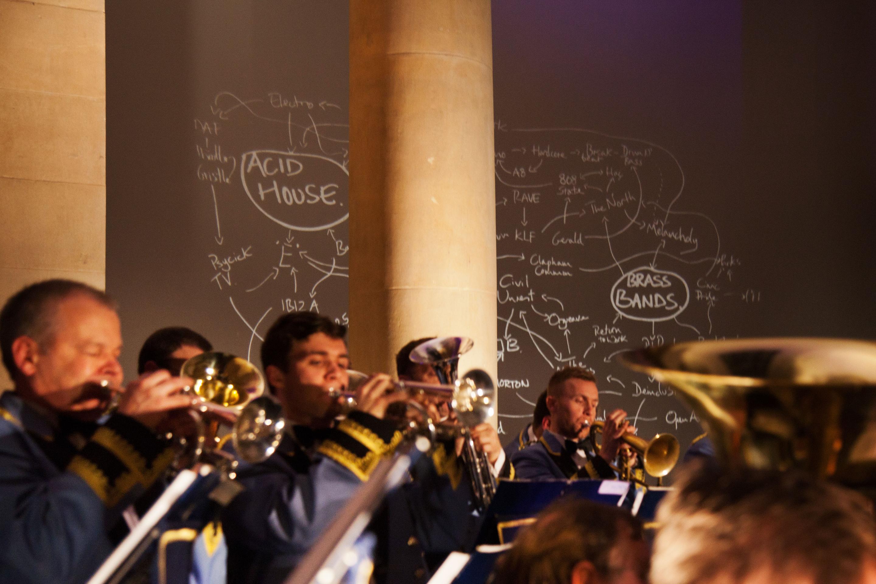 A brass band in uniform and playing trumpets and tubas performs in front of a mind map that connects acid house and brass band music.