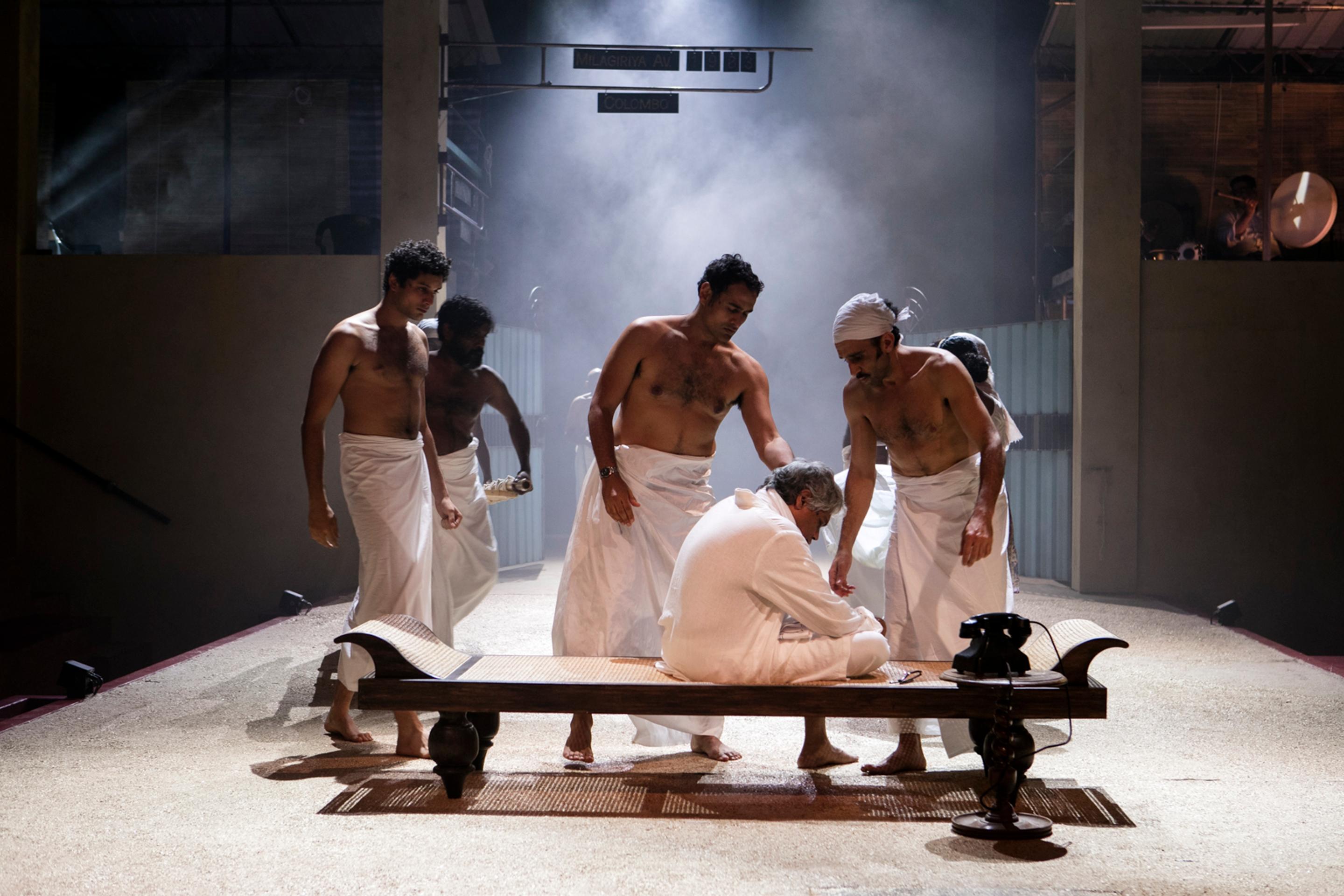 Men dressed in traditional white clothing and head wraps participate in a ceremonial activity around a long wooden bench, set against a smoky, atmospheric stage backdrop.