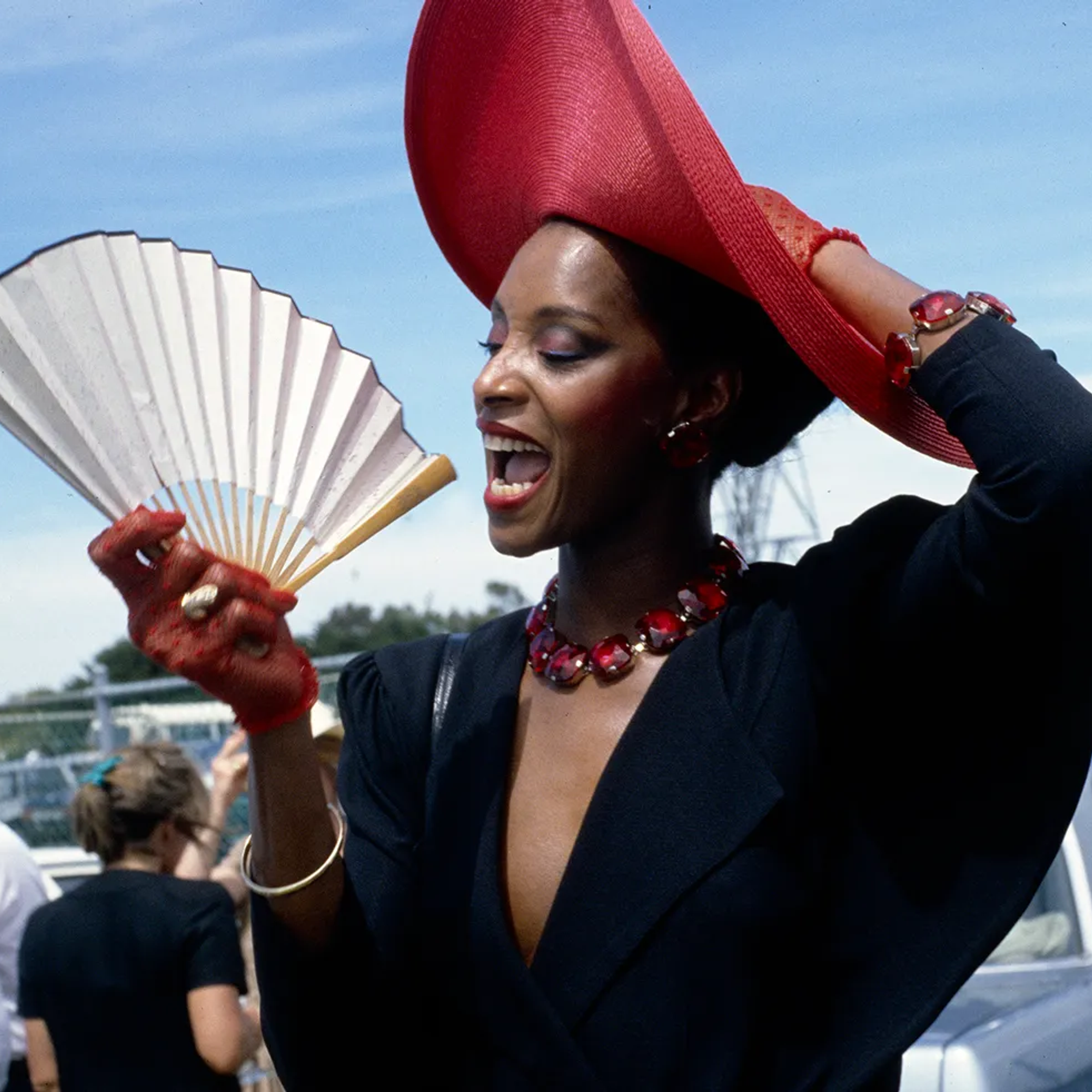 A woman poses holding onto her red fascinator-style hat with one hand and an unfurled fan in the other