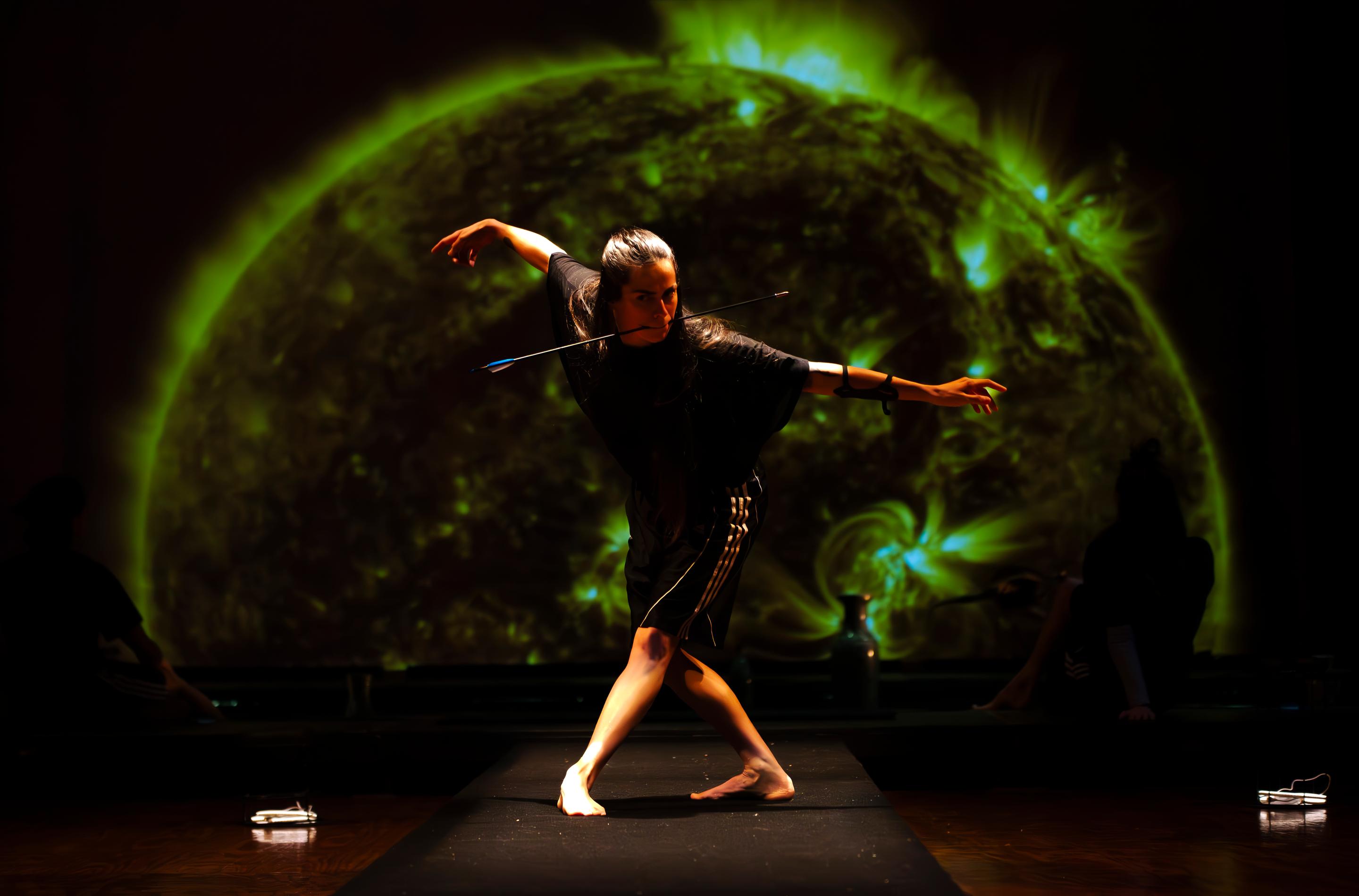 A dancer poses in front of a dark backdrop holding an arrow with her mouth.