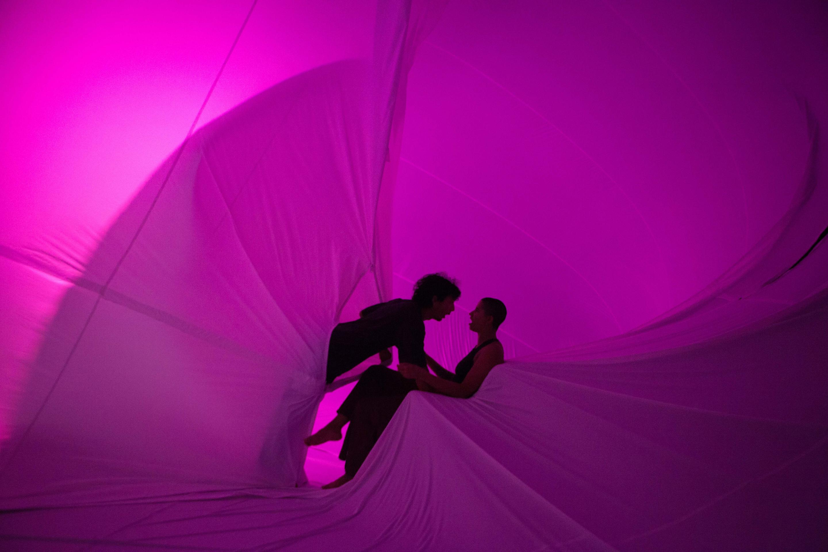 The silhouettes of two performers are pictured within an inflatable space. One is sitting, while the other leans over them carefully.