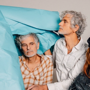 Three band members pose behind an enormous crinkled sheet of blue paper. 
