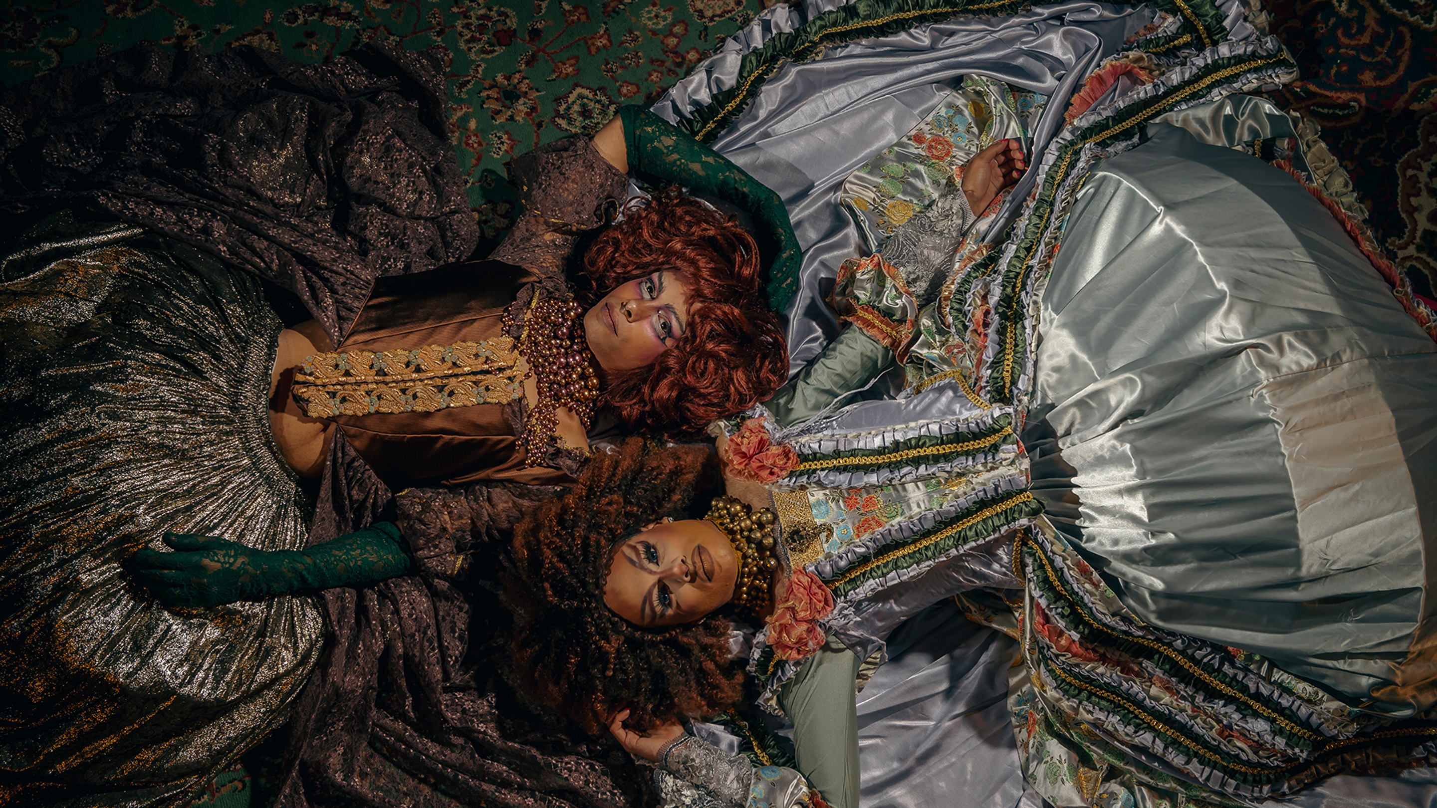 An aerial shot of two drag queens laying on their backs in full Elizabethan gowns and wigs