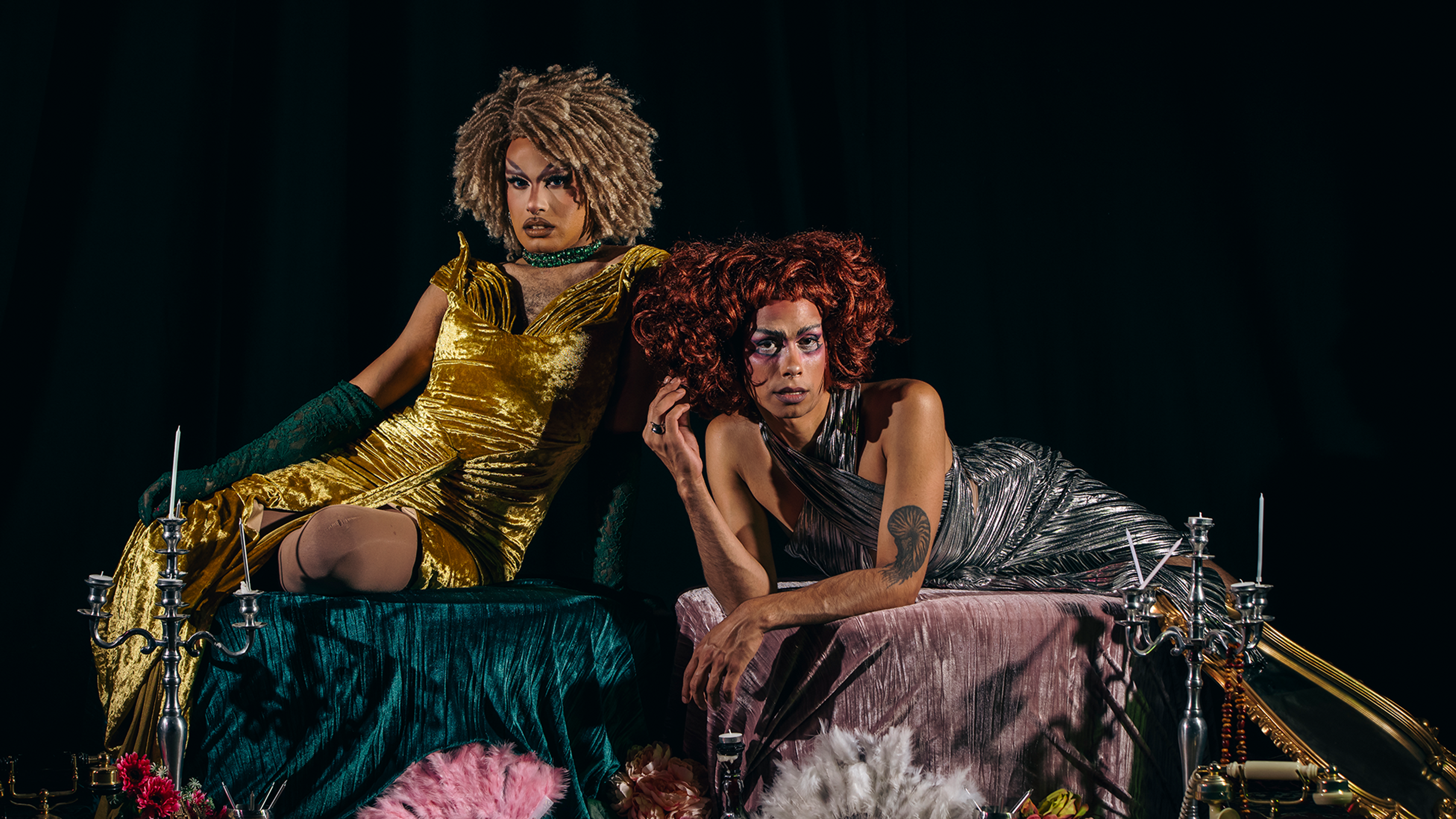 Drag queens Cerulean and Stone Motherless Cold recline back towards each other while wearing shiny, party dresses