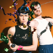 Two performers face the camera. One fires a confetti gun into the lens. 