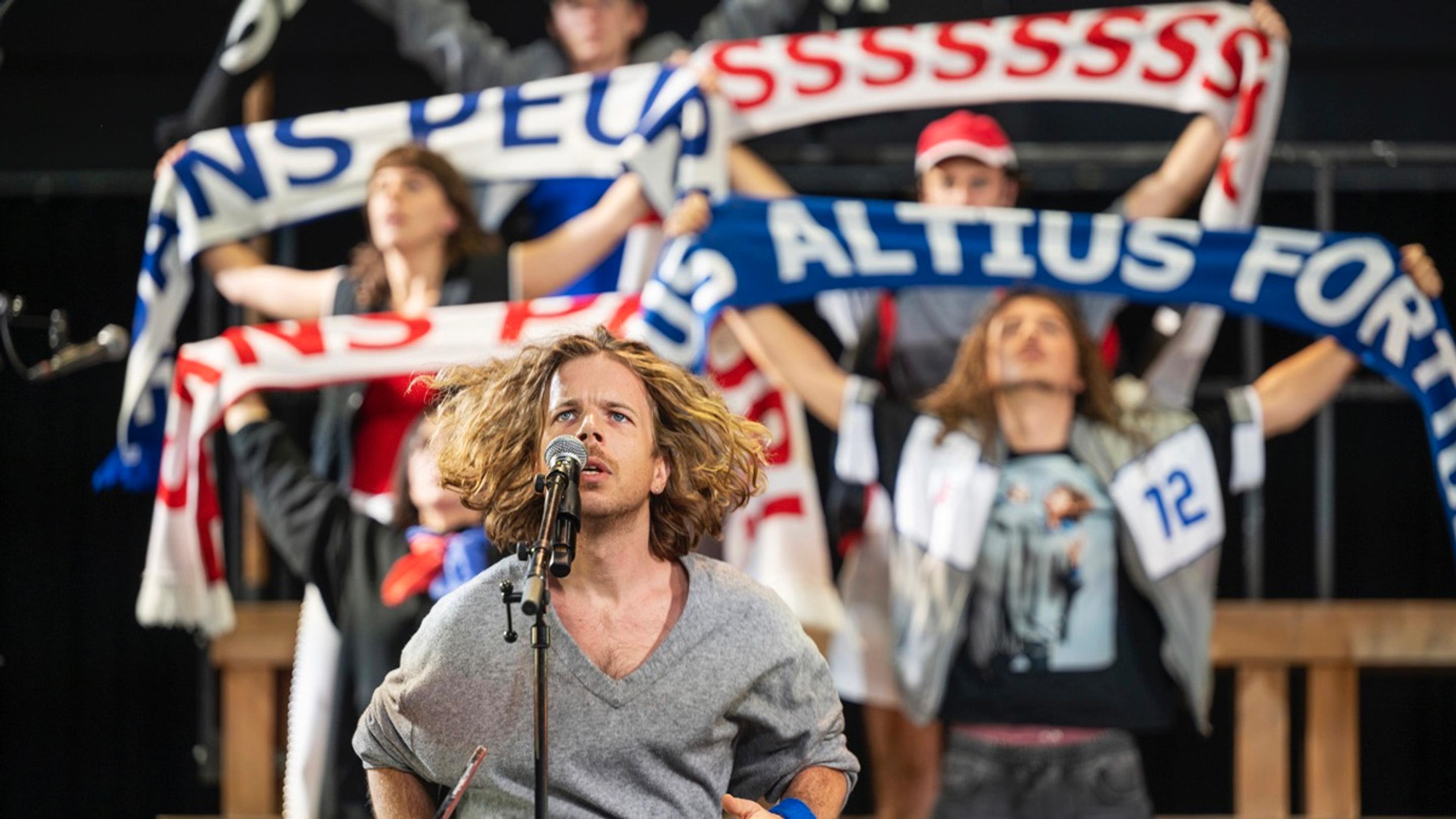 A performer stands in front of a microphone looking intently above the camera. Behind them, members of an ensemble stand on a grandstand holding scarves for the audience to see.