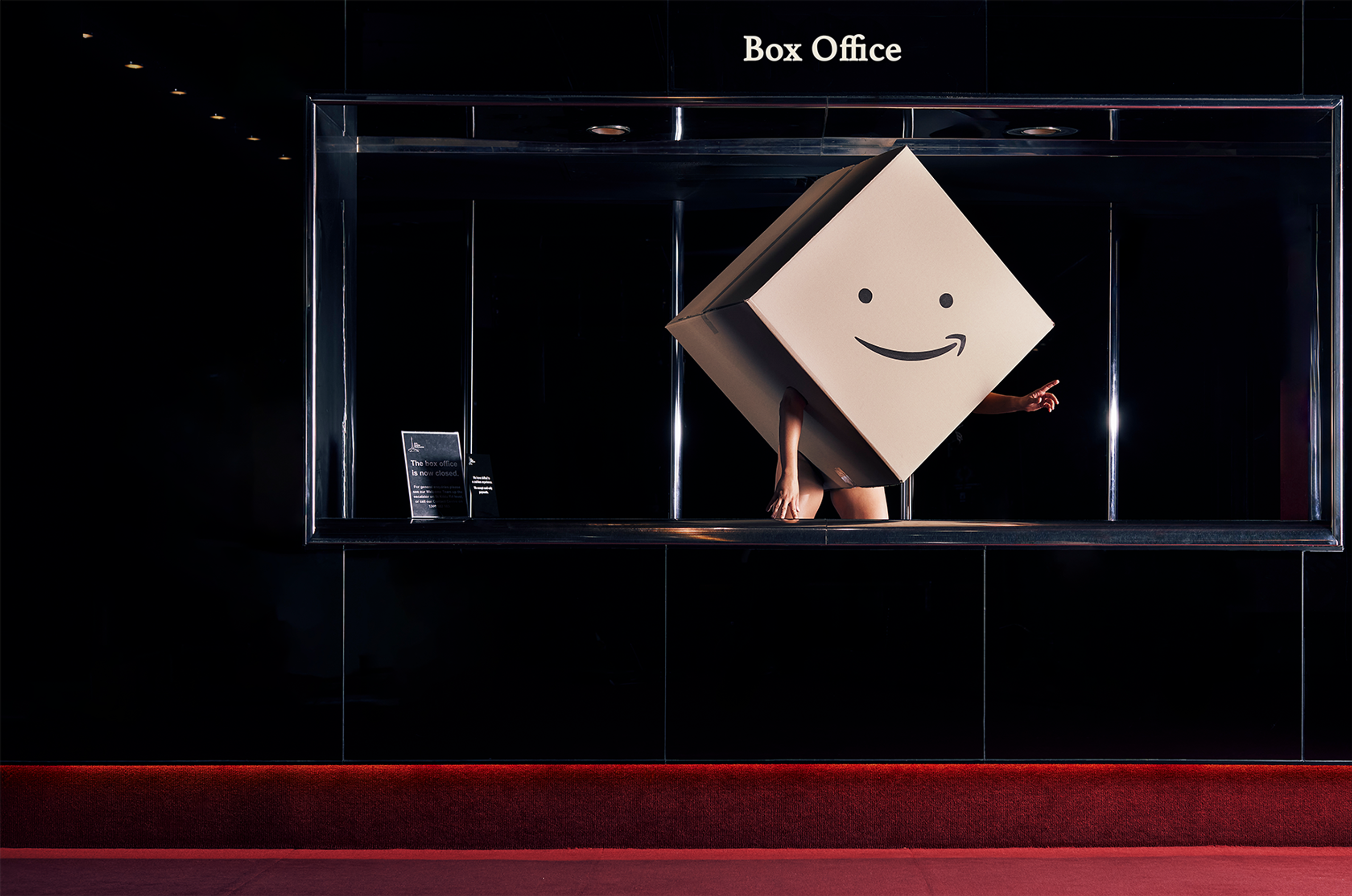 A person wearing a giant box, printed with a smile on the front, stands in a dim Box Office. 