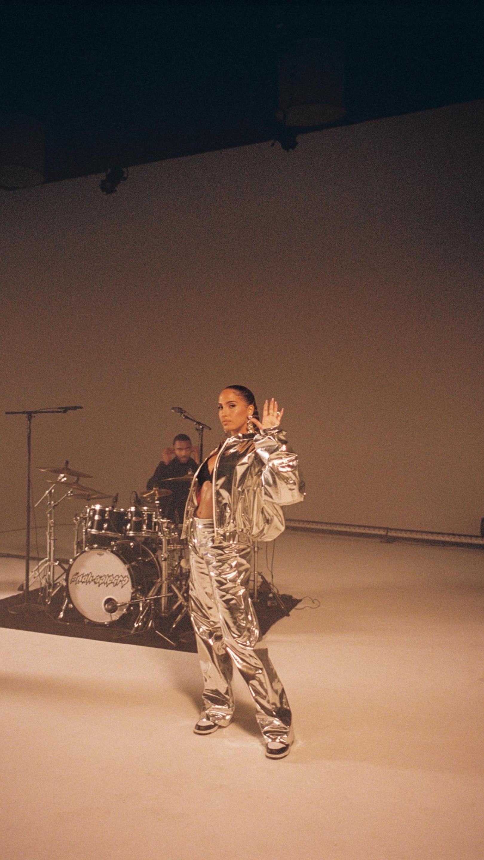 A musician wearing a reflective silver suit looks at the camera and waves and a person plays a drum kit behind them.