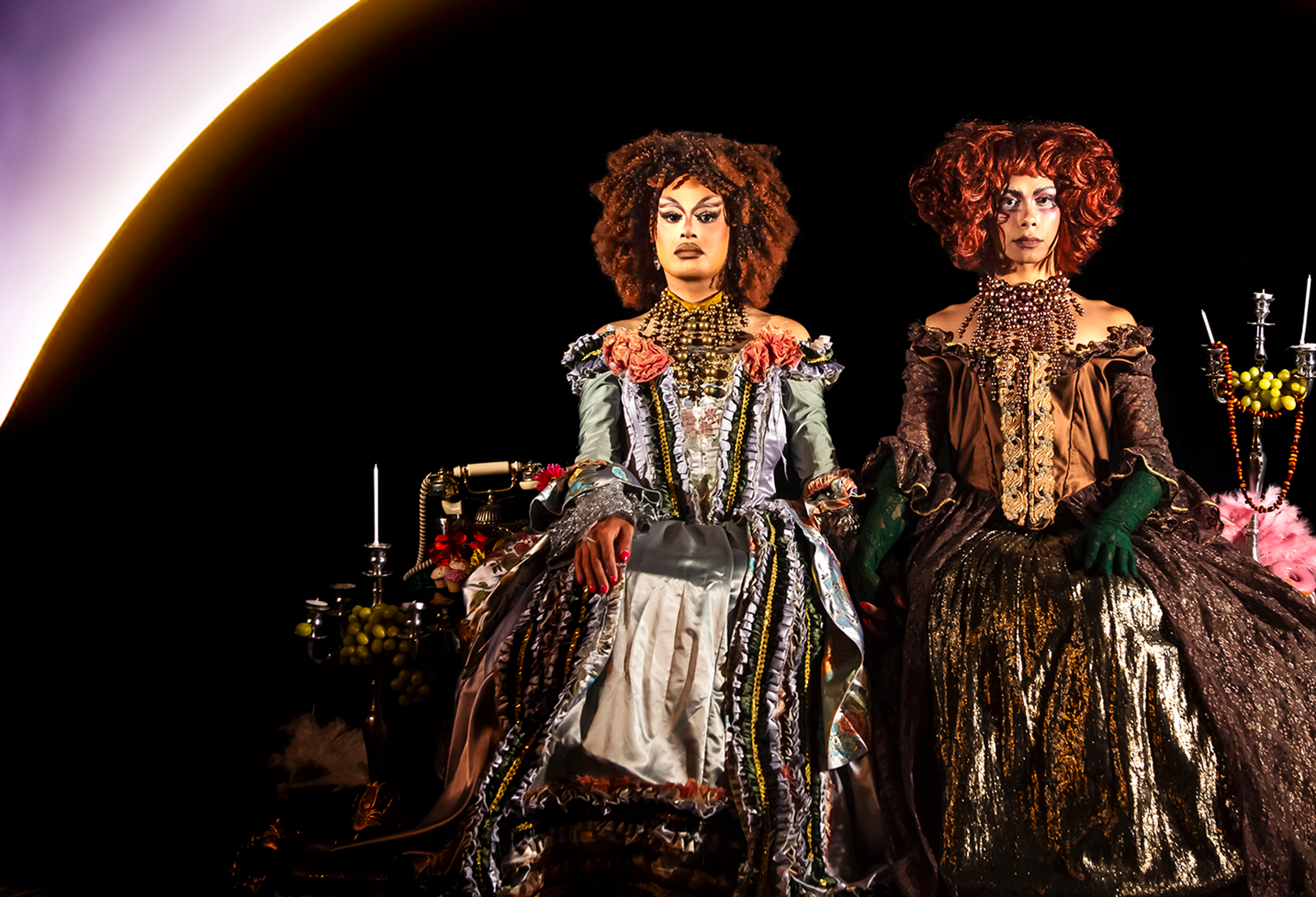 Two drag queens dressed in regal attire sit side by side, as if posing for a royal portrait. 