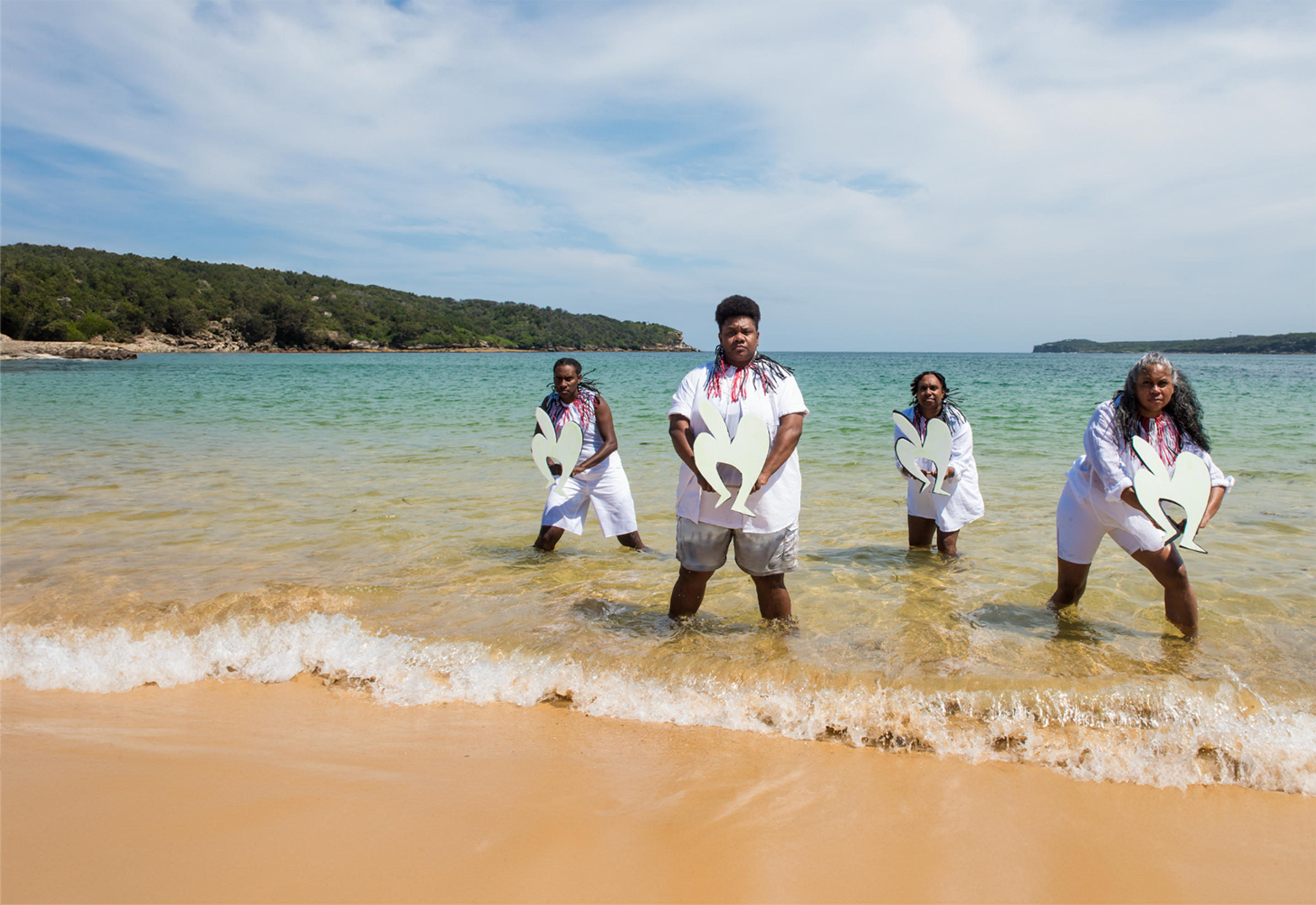A group of people stand in the shallows of a tropical beach, holding cut-out shapes. 