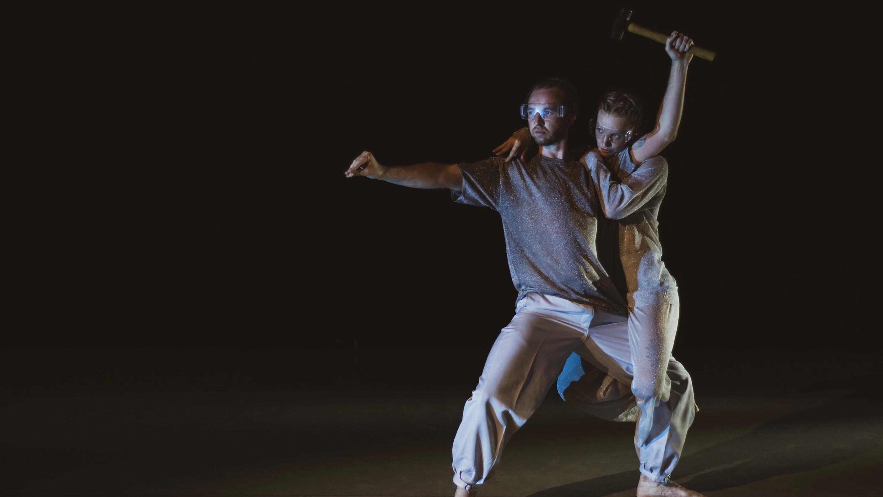Two dancers embrace in a fighting position, one holding a hammer above their heads.