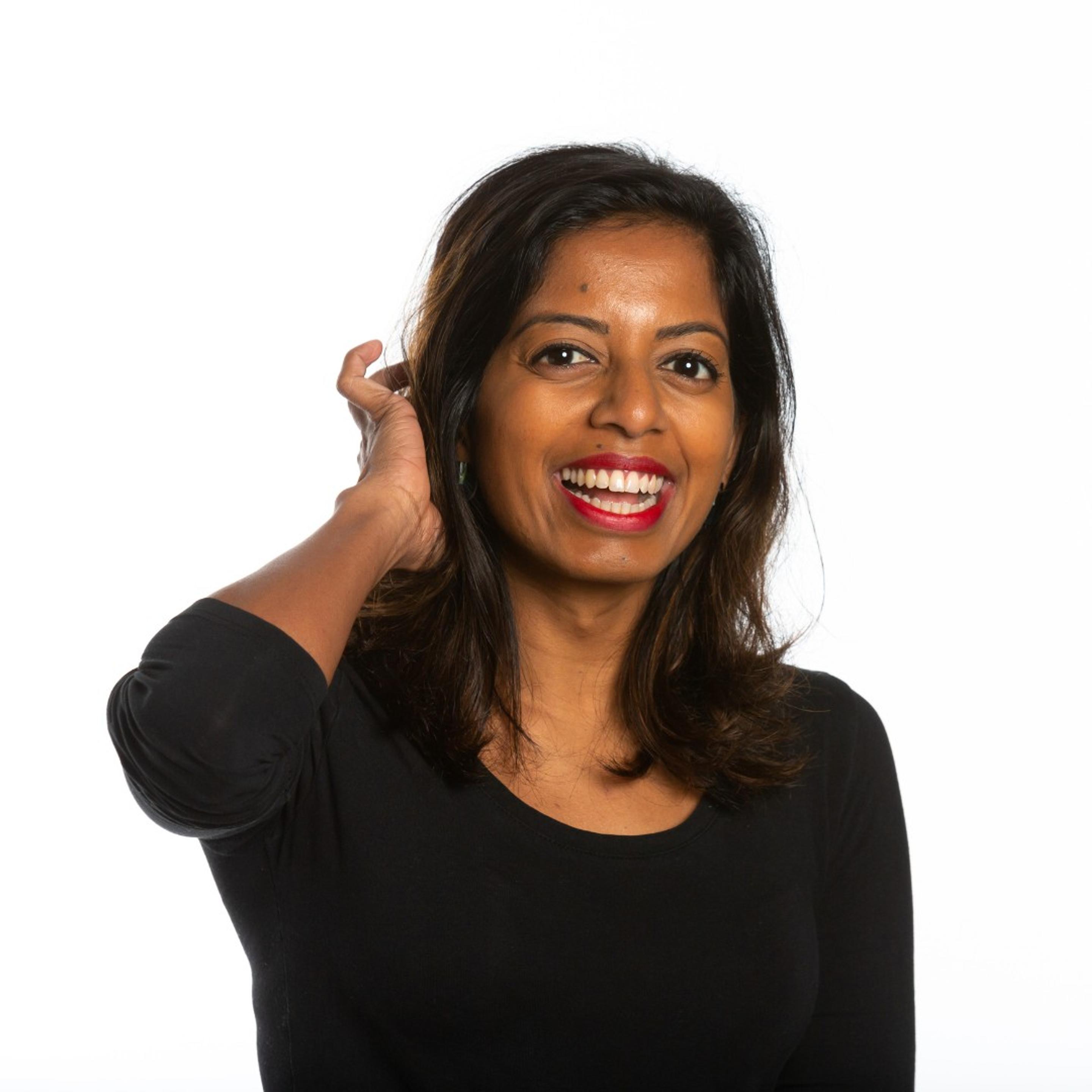 Comedian Sashi Perera smiles at the camera while tucking her hair behind her ear