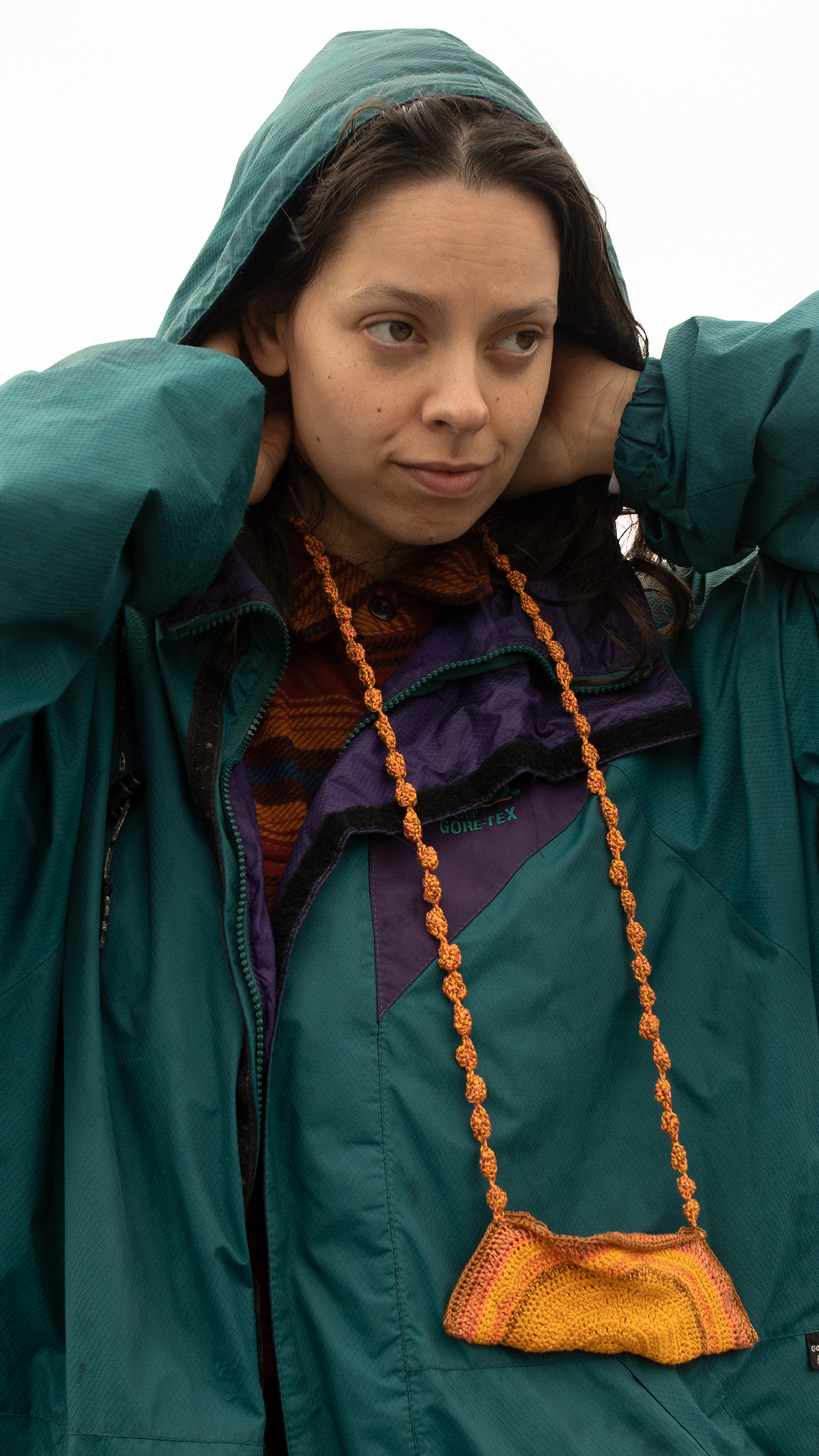 A person in an oversized parker and hood on their head, looking out of right of frame, their hands are reaching inside the hood to their neck.