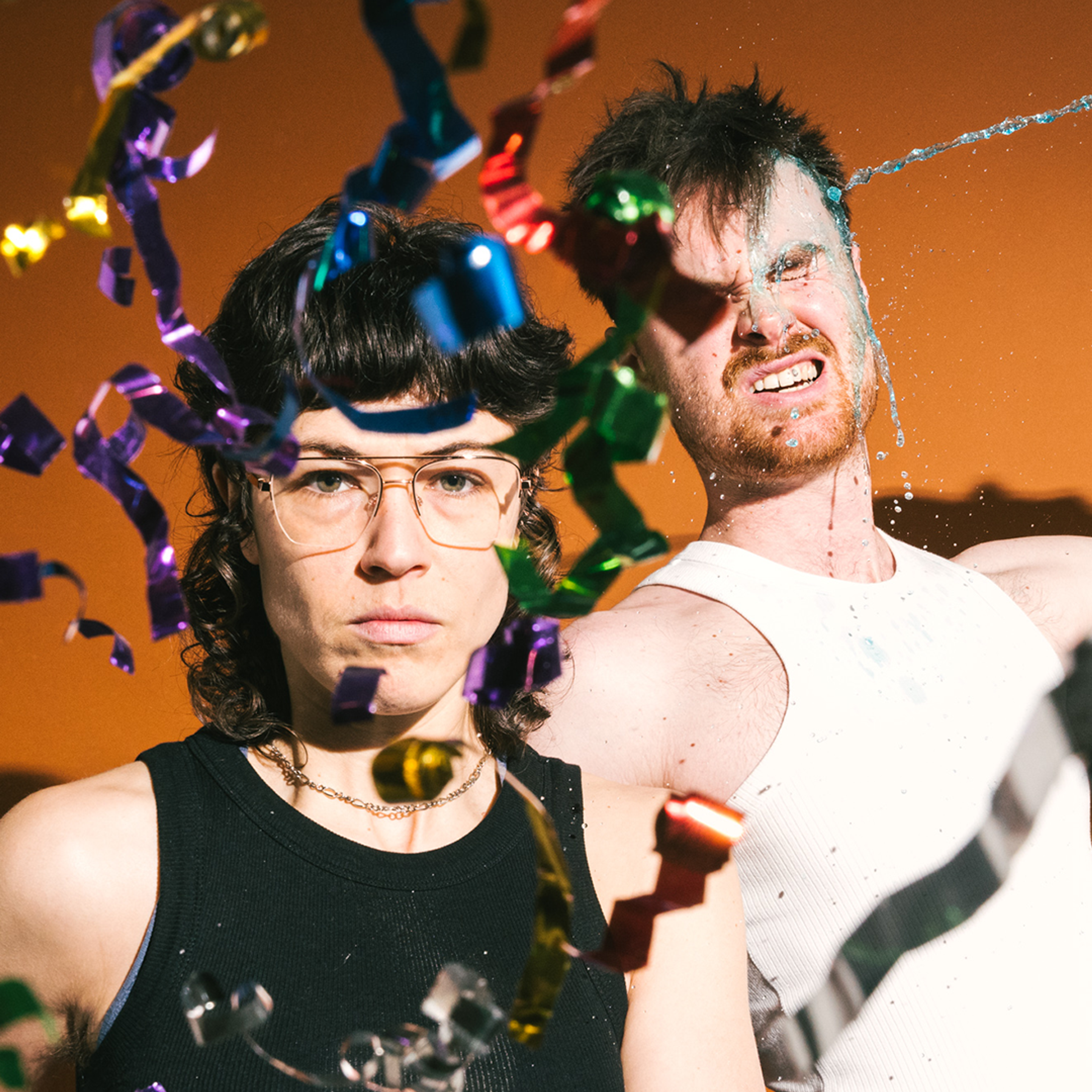 Two performers face the camera. One fires a confetti gun into the lens. 