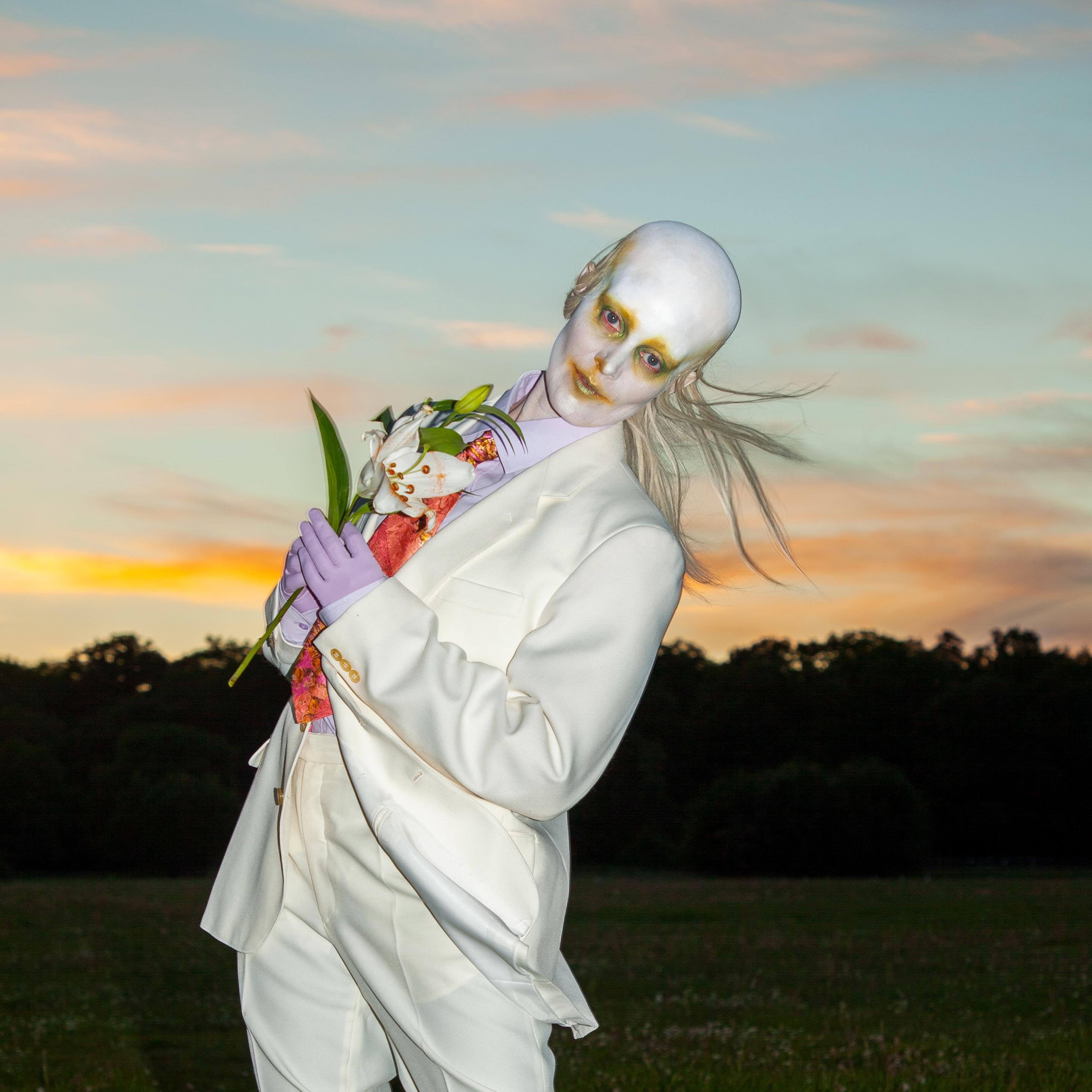 Karin Dreijer aka Fever Ray wears a bald cap and all white makeup, and a traditional masc white suit, holding a flower posey 