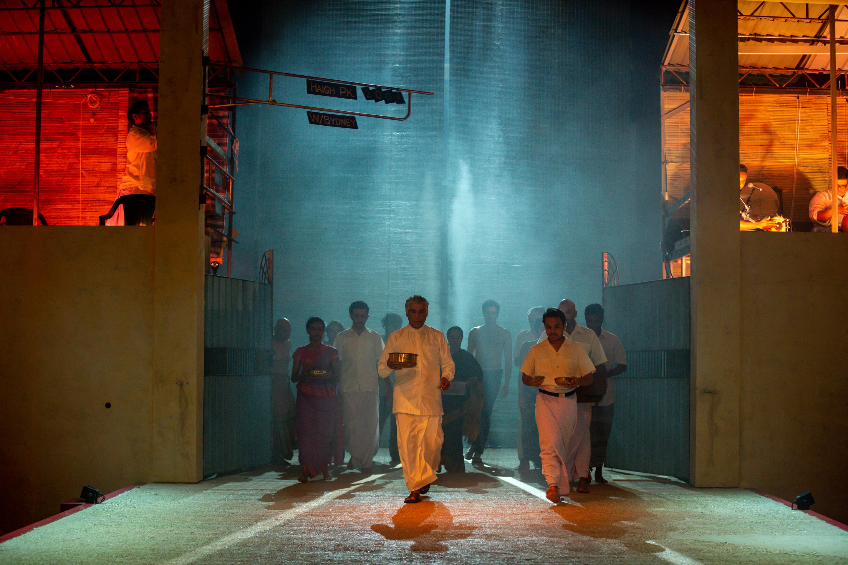 A procession of people dressed in white traditional clothes emerges from a beam of light in a misty, atmospheric setting.