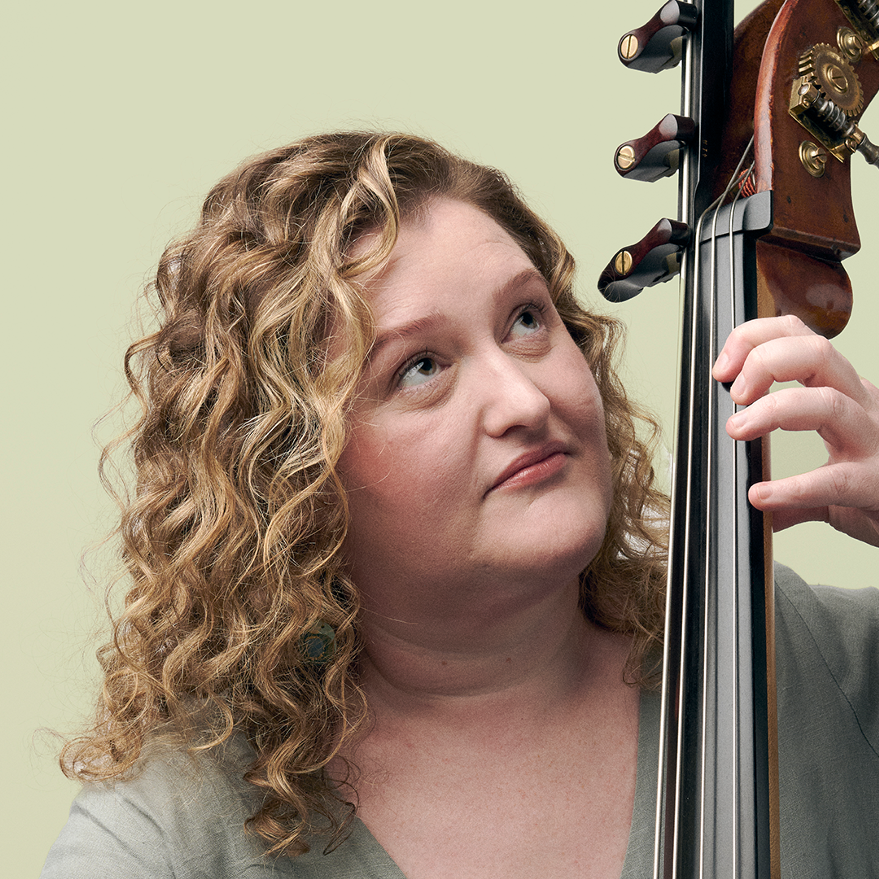A woman with curly hair stands holding her cello, looking upwards