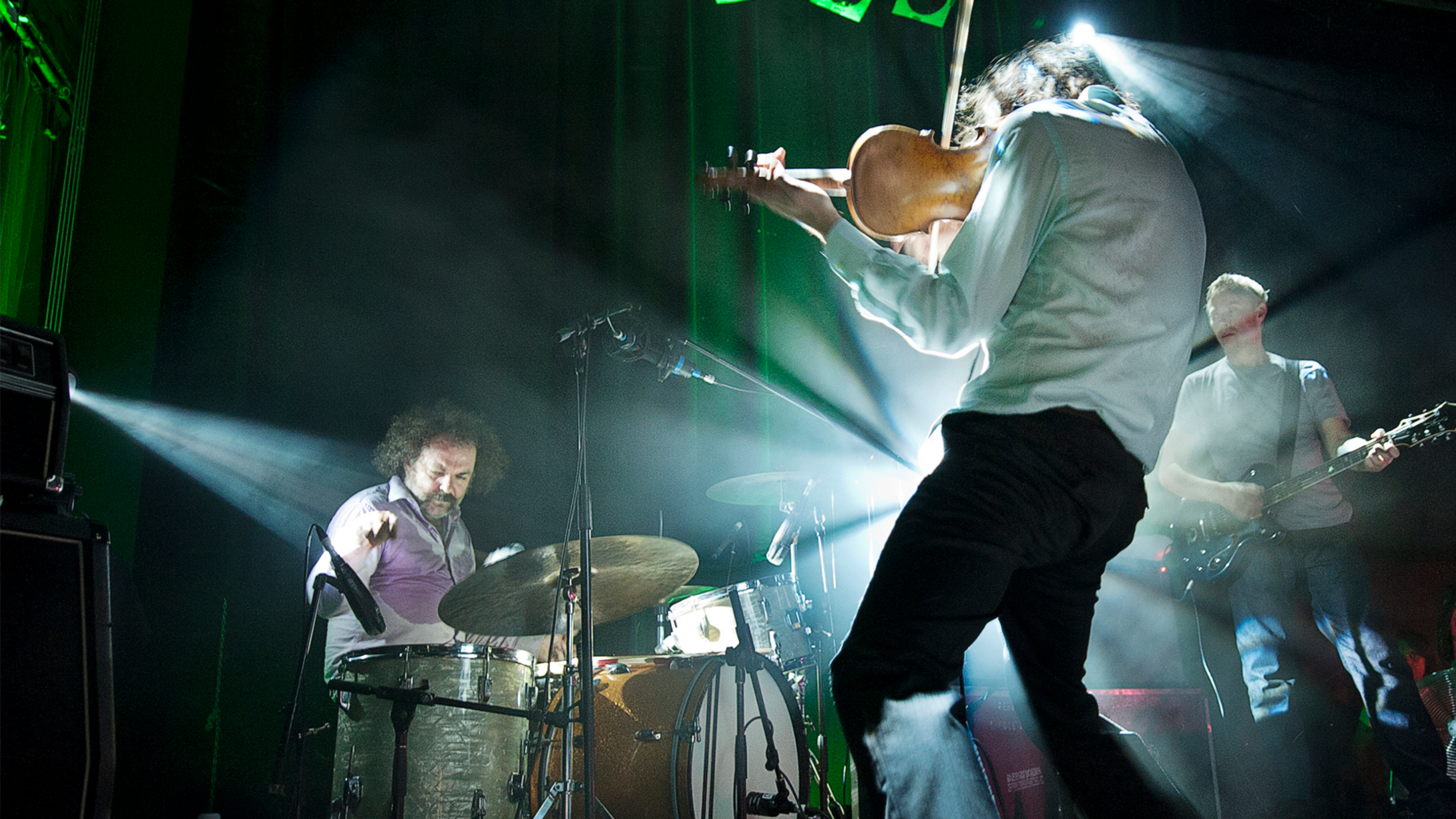A dynamic live music scene with a violinist in motion, a drummer, and a bassist performing on stage under bright lights, capturing a moment of artistic expression.