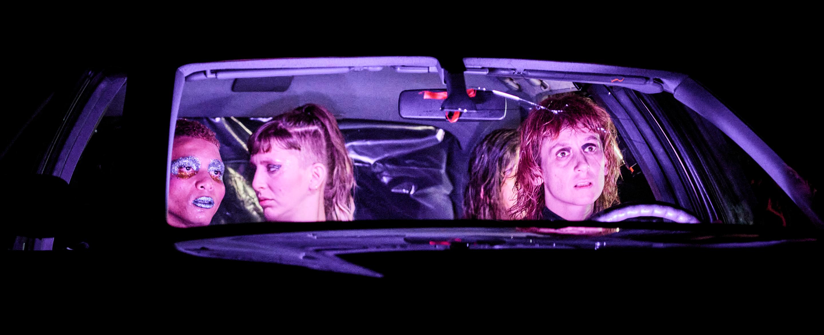 Four people sit in a car, their faces lit up by neon light. They all look out of the car in different directions. 