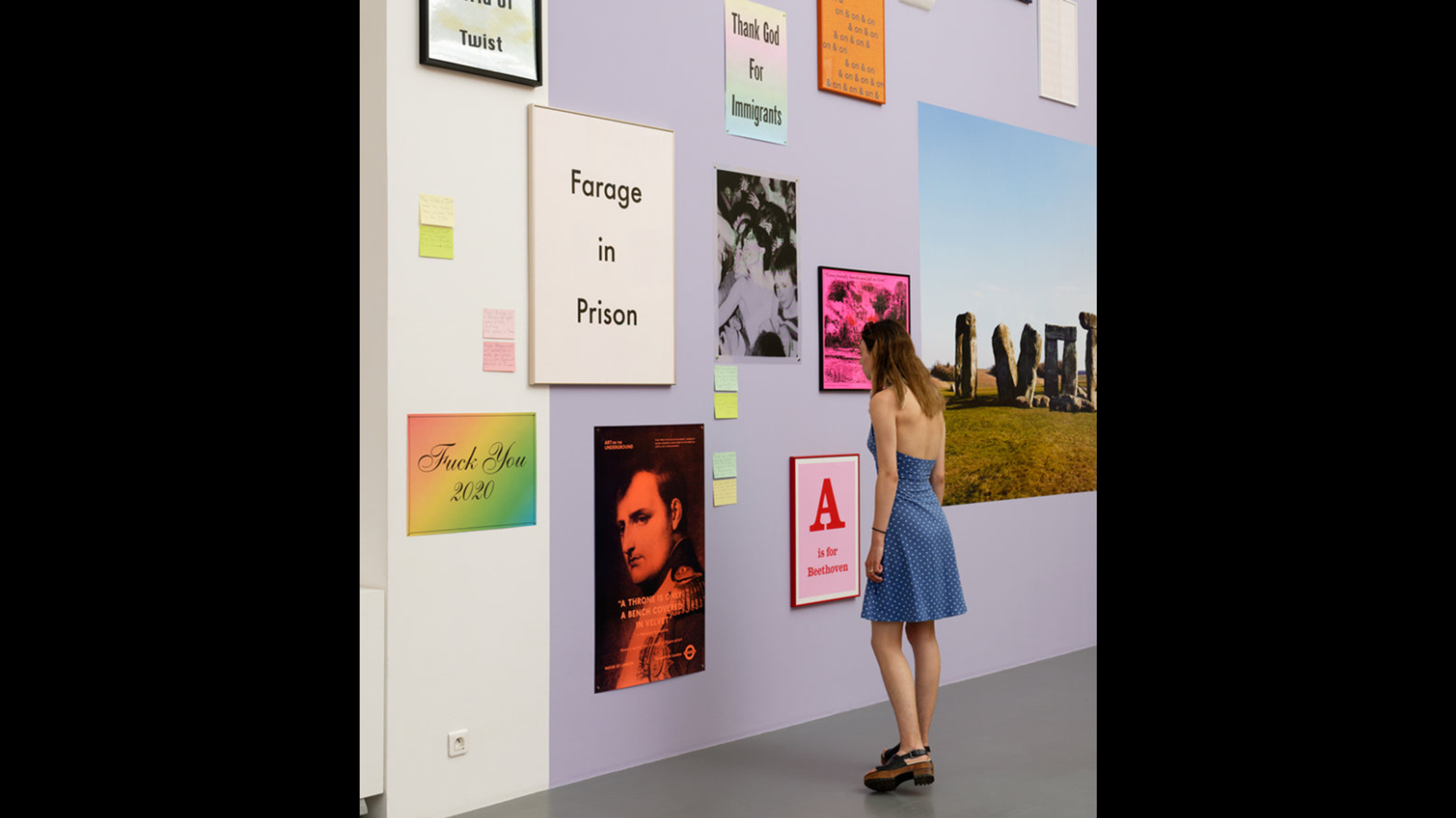 A person in a dress stands looking at poster artwork by Jeremy Deller