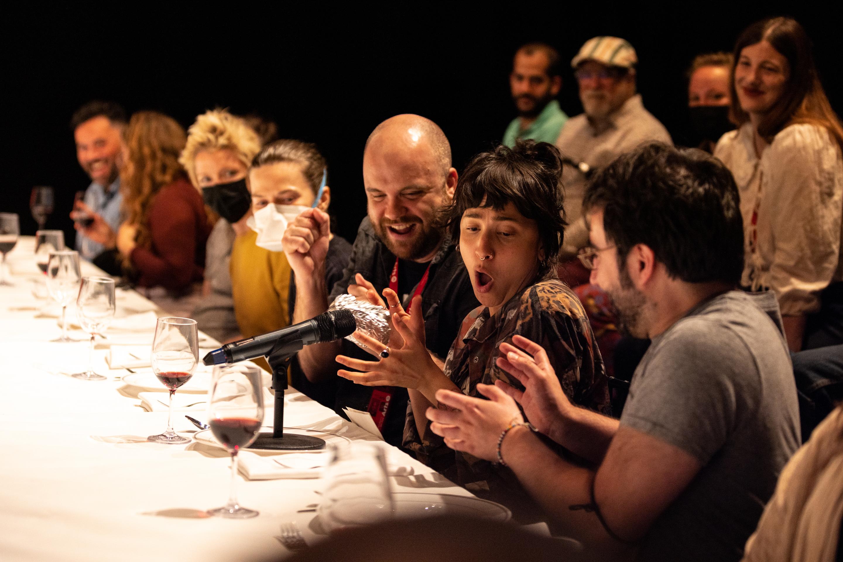 An audience sits in front of a dinner table with plates and wine glasses in front of them. A microphone is on the table and the audience are interacting with it animatedly.