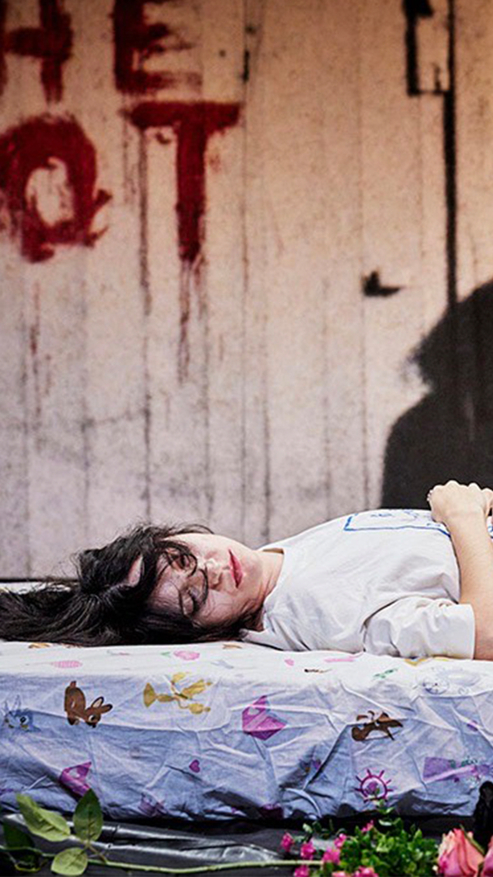 A woman lies sleeping on a mattress surrounded by roses. Her face turned to the camera. "She Got Love" is scrawled on the wall behind her in red paint.
