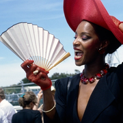 A woman poses holding onto her red fascinator-style hat with one hand and an unfurled fan in the other