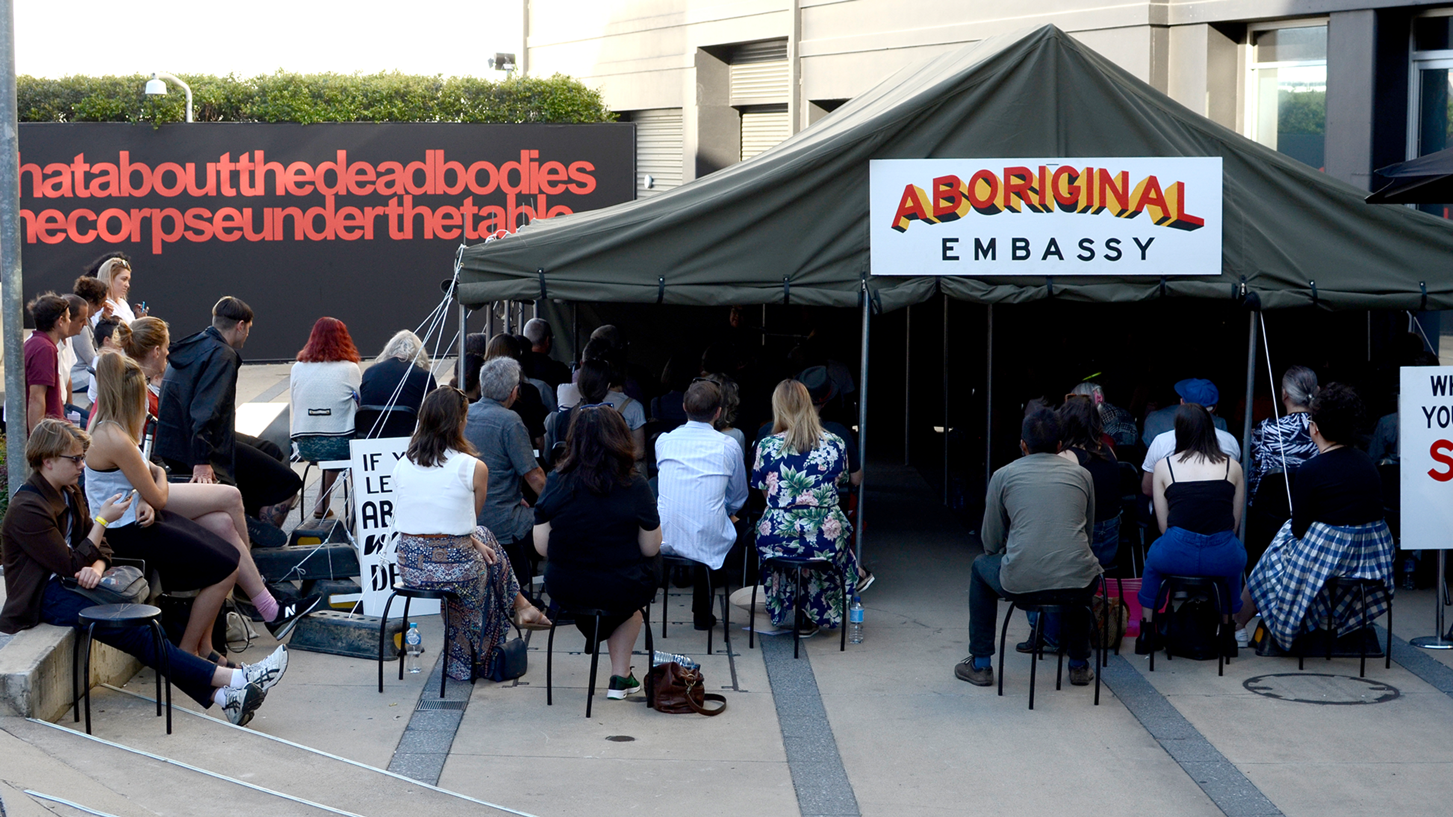 A large canvas tent with the sign 'Aboriginal Embassy'. People seated on stools gather around its entrance listening to a speaker 