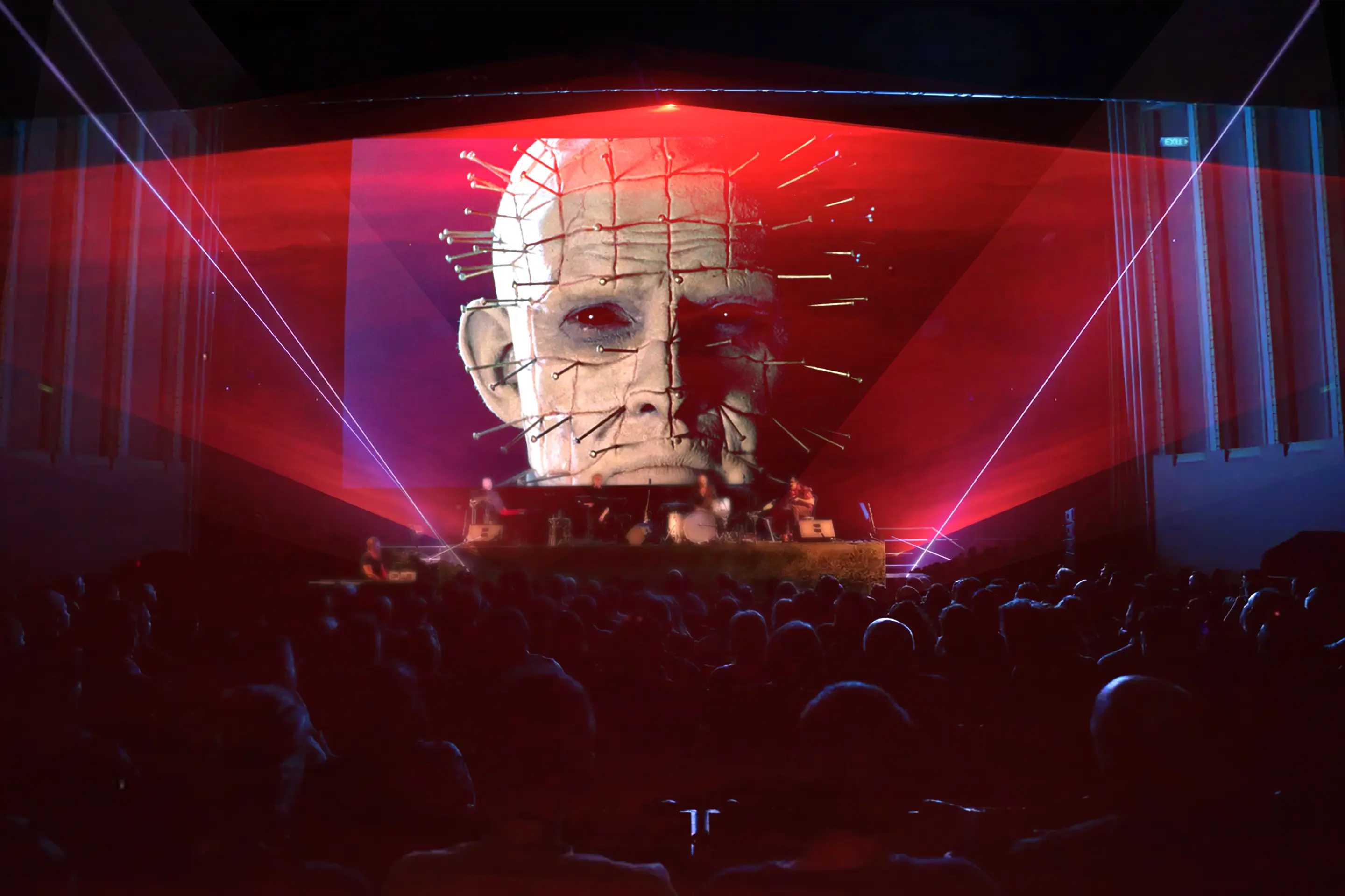 A theatre full of people watching a film screen featuring a still of Hellraiser. A band performs on stage and lasers light up in front of the screen.