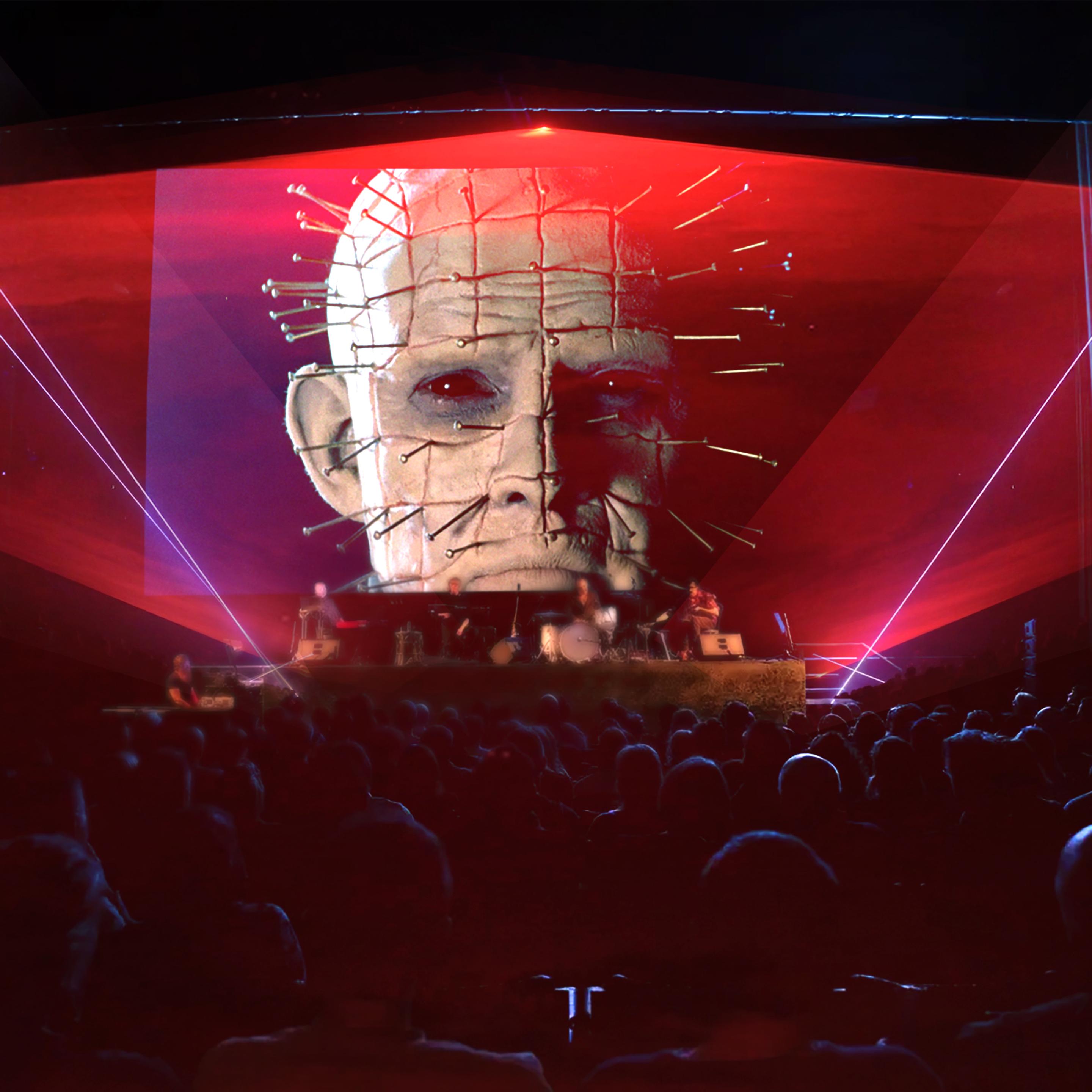 A theatre full of people watching a film screen featuring a still of Hellraiser. A band performs on stage and lasers light up in front of the screen.