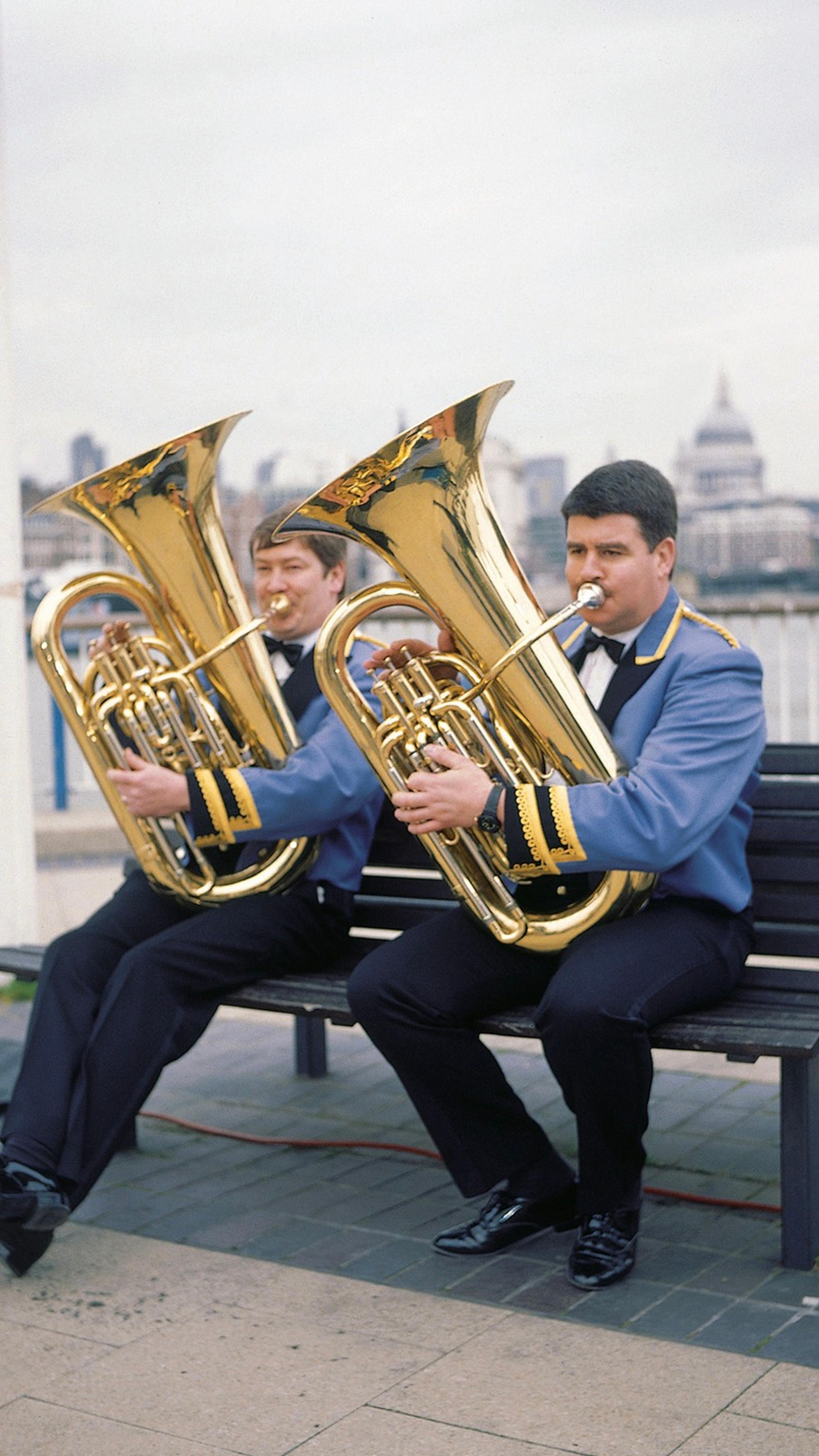 Two men in matching uniforms sit outside on a bench, each playing a tuba