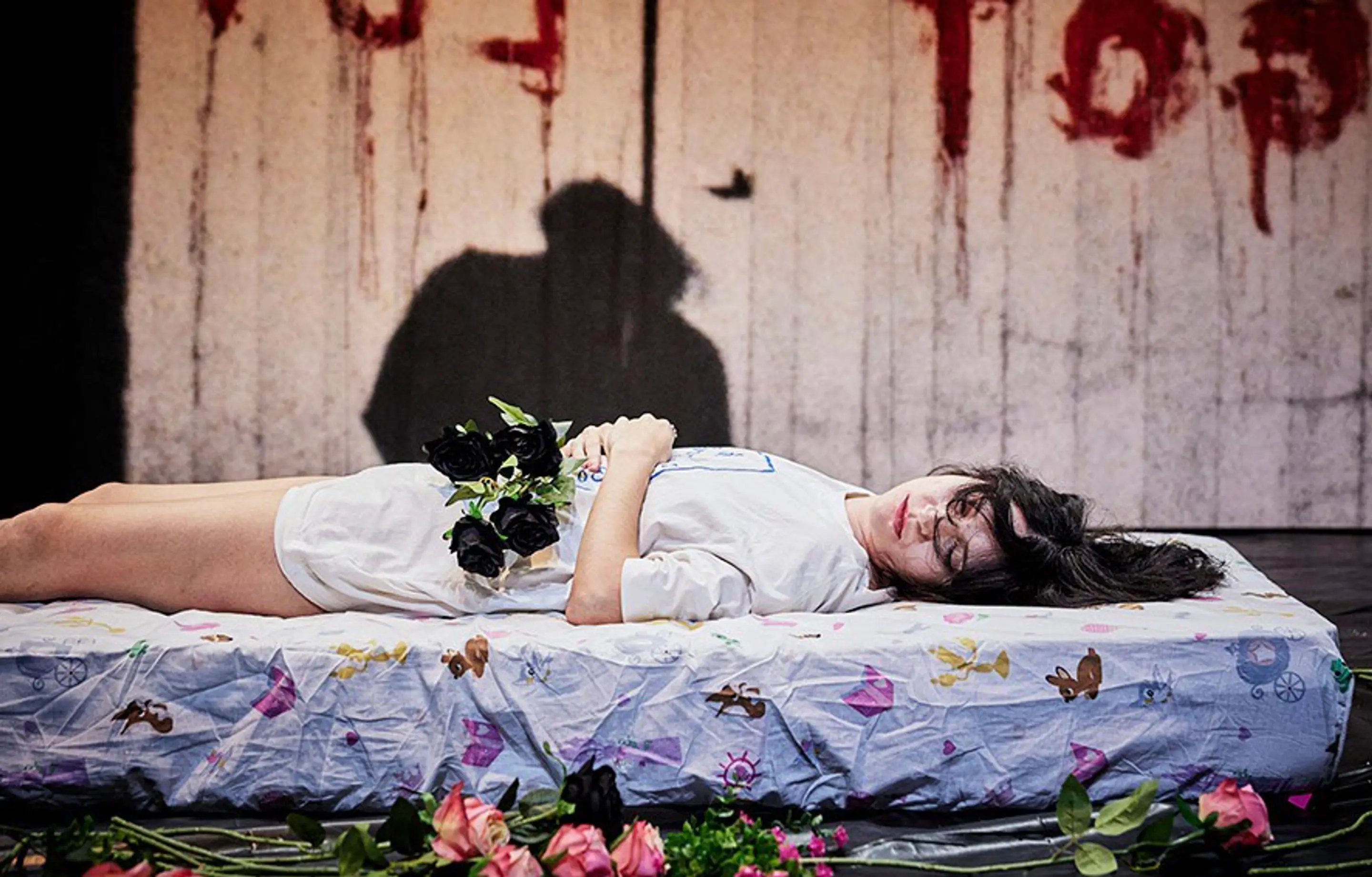 A woman lies sleeping on a mattress surrounded by roses. Her face turned to the camera. "She Got Love" is scrawled on the wall behind her in red paint.