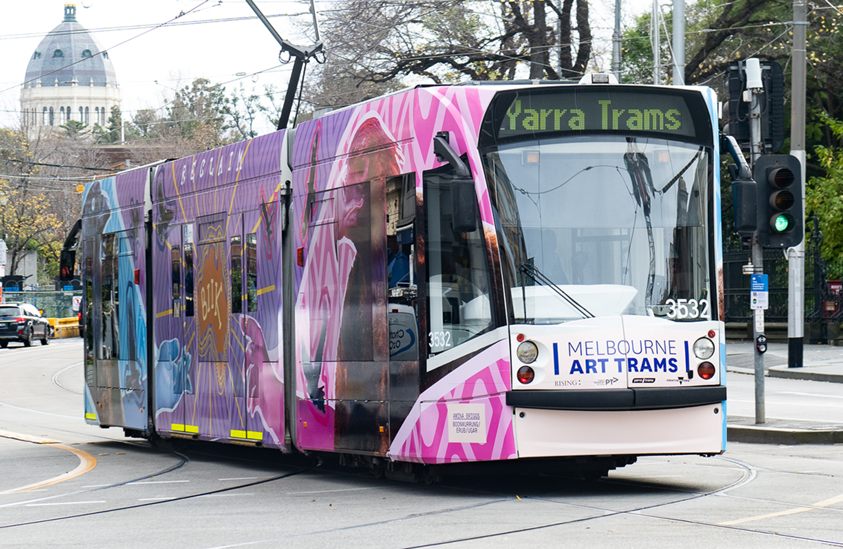 Amina Briggs' Art tram driving down Spring Street, alongside Parliament House Melbourne, with city buildings in the backdrop.
