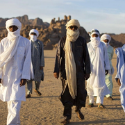 A group of men wearing white and blue robes and head coverings, in the desert.