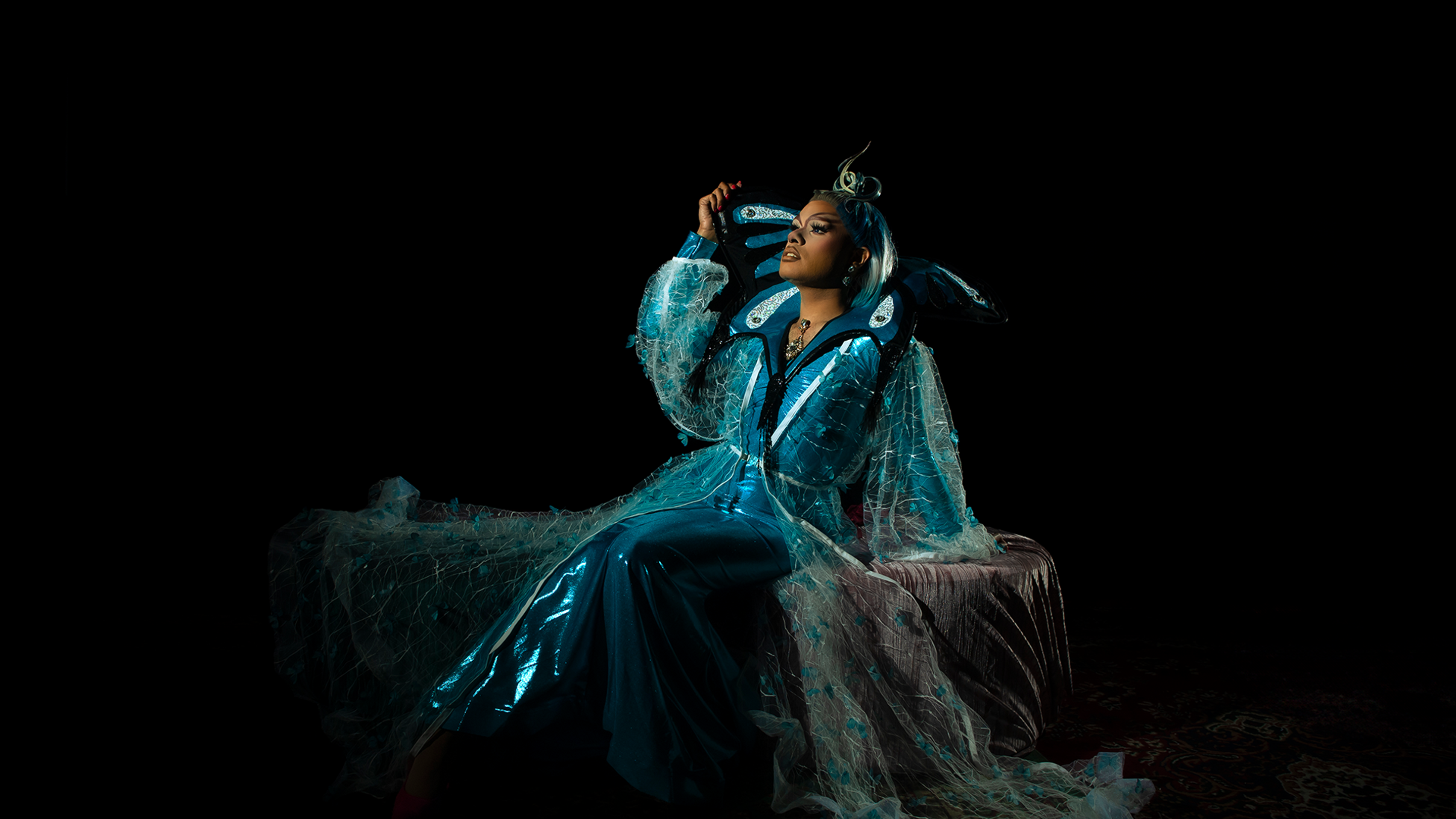 Cerulean, a First Nations drag queen in flowing royal dress leans back in their seat, looking up toward the top left of the frame