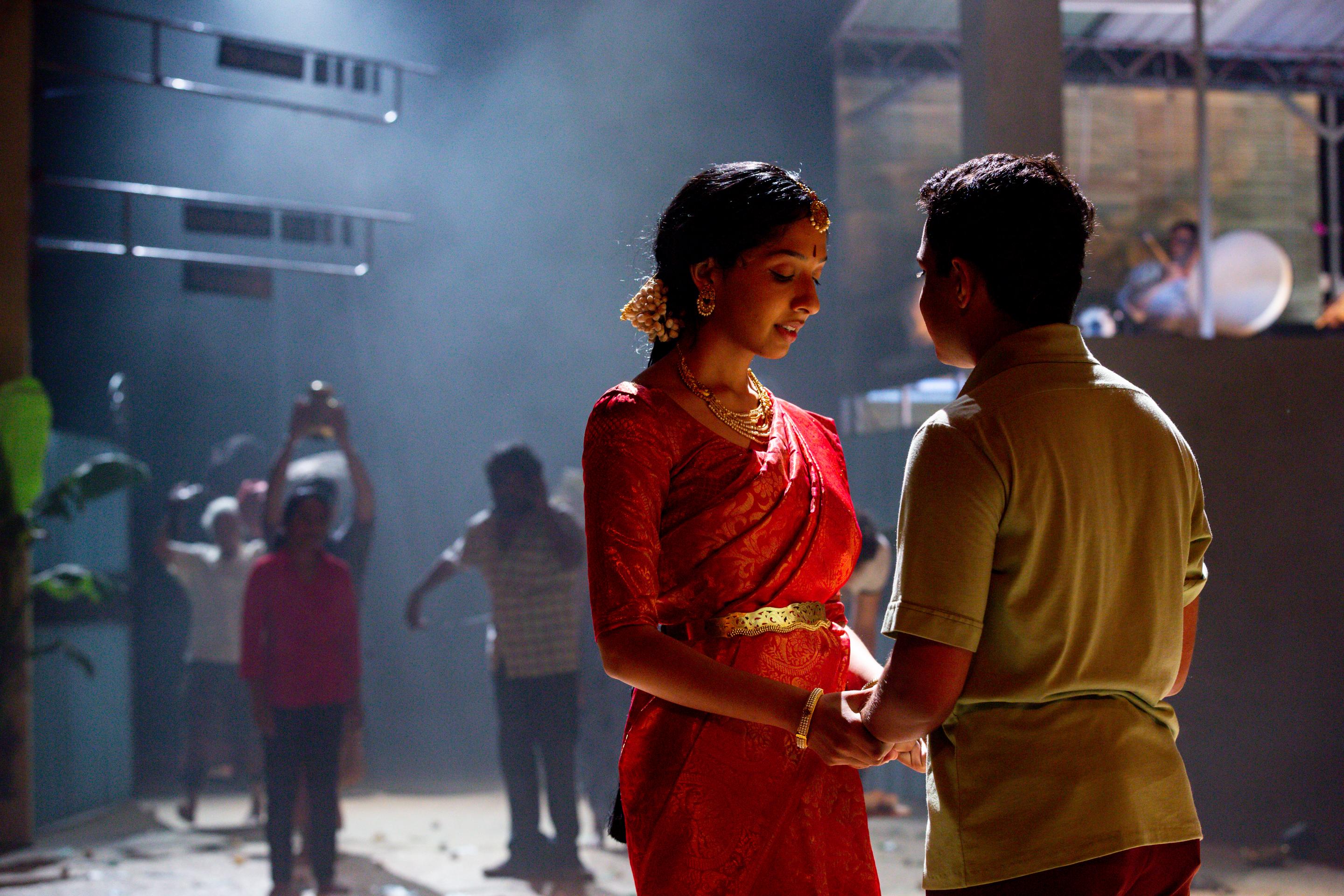 A woman in a red saree and a man in a tan shirt hold hands, sharing an intimate moment on a dimly lit stage, with the crew working in the background.
