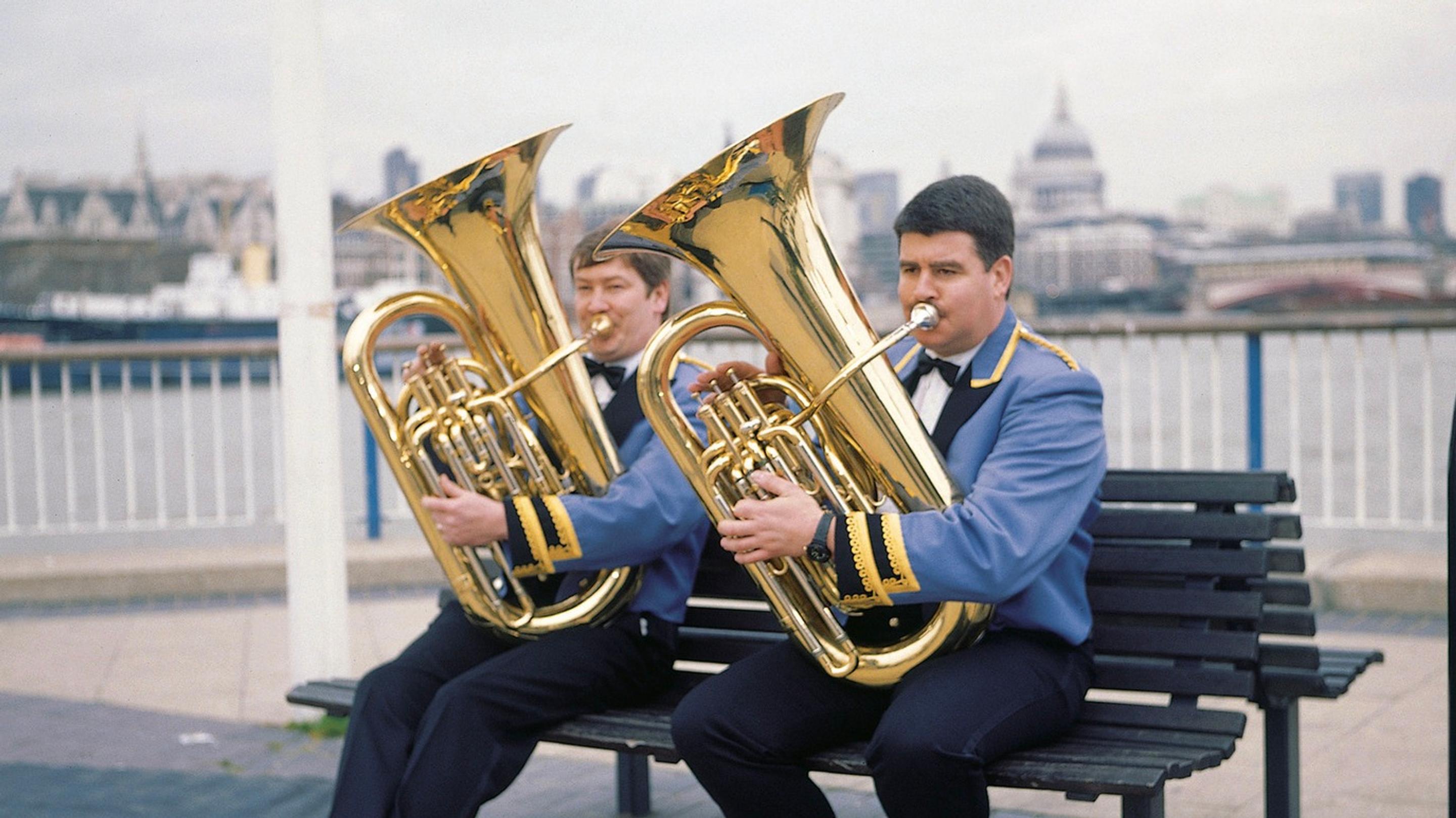 Two men in matching uniforms sit outside on a bench, each playing a tuba
