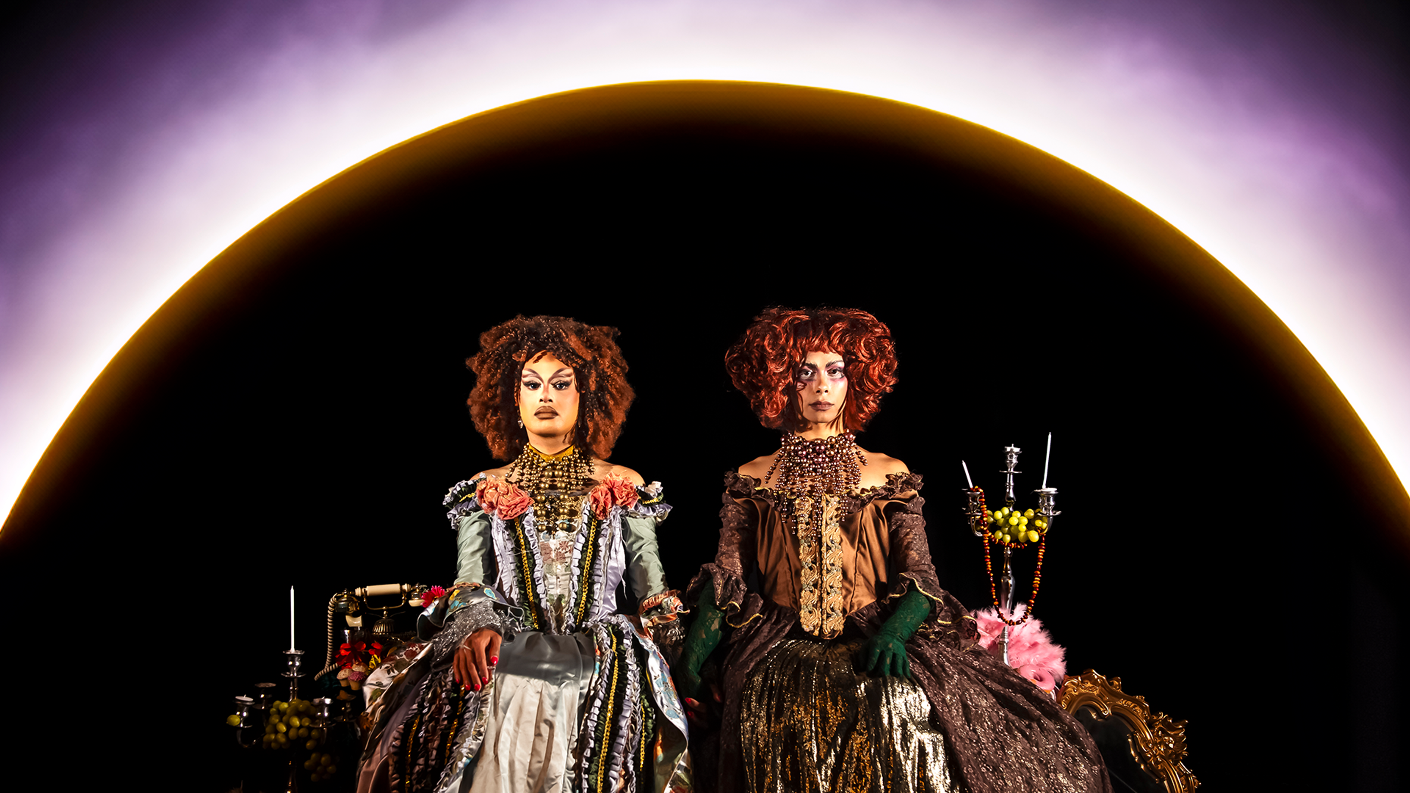 Two drag queens dressed in regal attire sit side by side, as if posing for a royal portrait. 