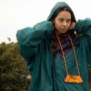 A person in an oversized parker and hood on their head, looking out of right of frame, their hands are reaching inside the hood to their neck.