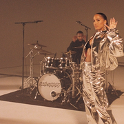 A musician wearing a reflective silver suit looks at the camera and waves and a person plays a drum kit behind them.